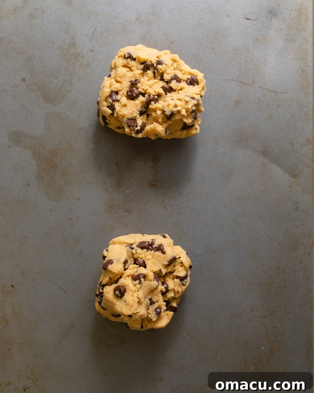 Two large, irregularly shaped cookie dough balls placed on a parchment-lined baking sheet, ready for the oven.
