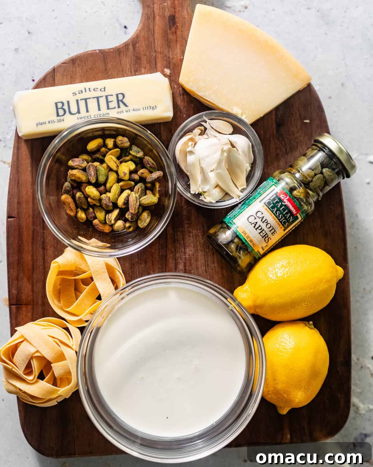 Arrangement of fresh ingredients for lemon caper pasta: Parmesan cheese, fresh lemons, capers, pistachios, dry pasta, butter, olive oil, fresh parsley, and garlic cloves.