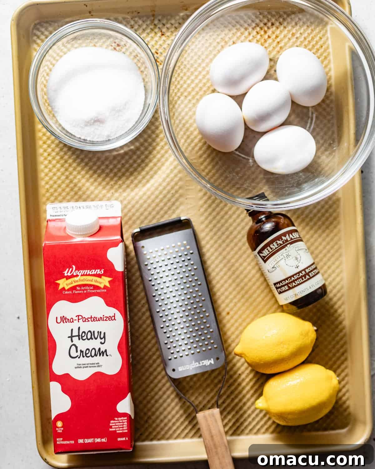 Carefully arranged ingredients for lemon creme brulee on a pristine baking sheet: heavy cream, whole milk, separated egg yolks, fresh lemons, vanilla bean paste, granulated sugar, and lemon extract.