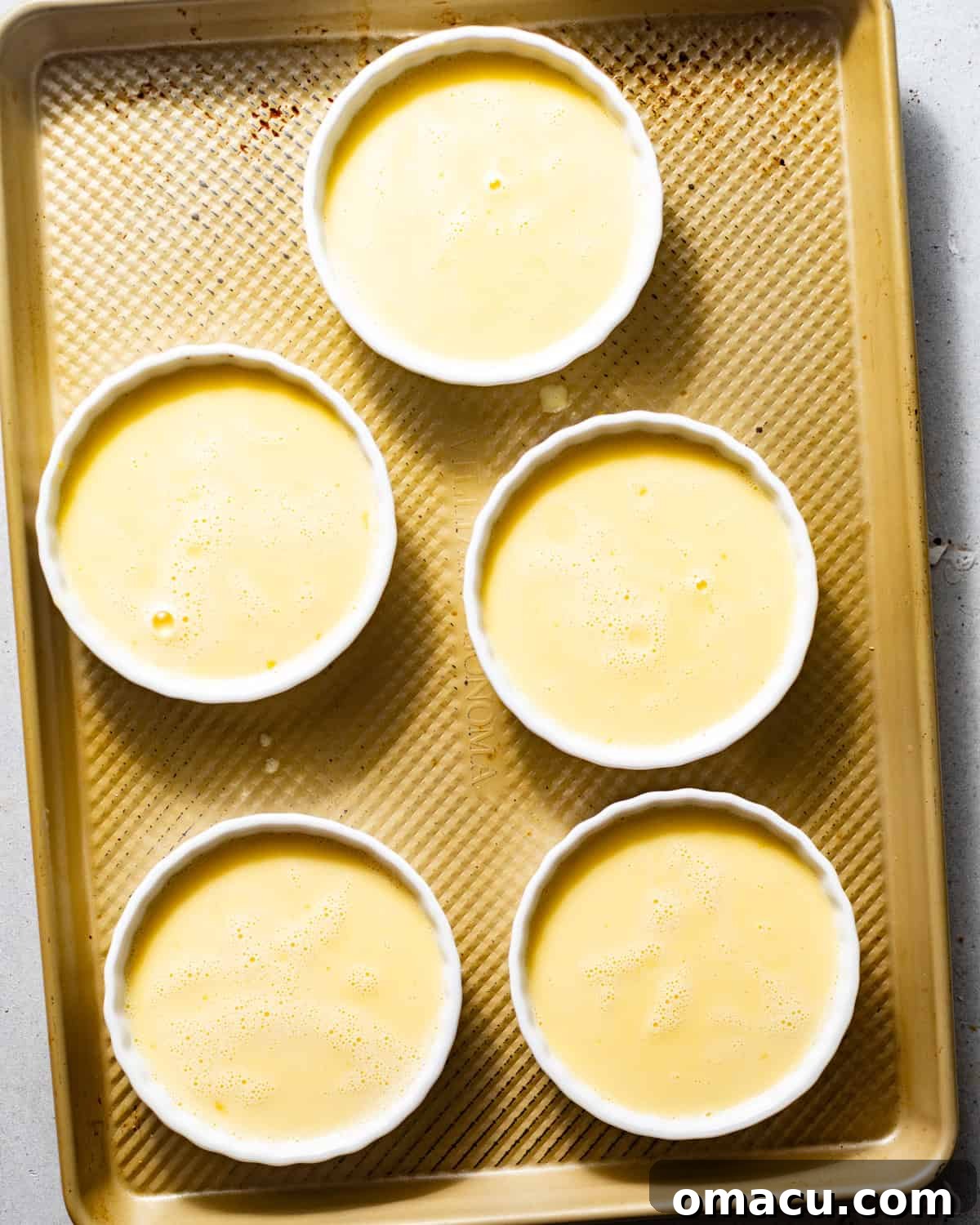 Individual ramekins filled with lemon creme brulee custard on a baking sheet, surrounded by hot water for a gentle water bath during baking.