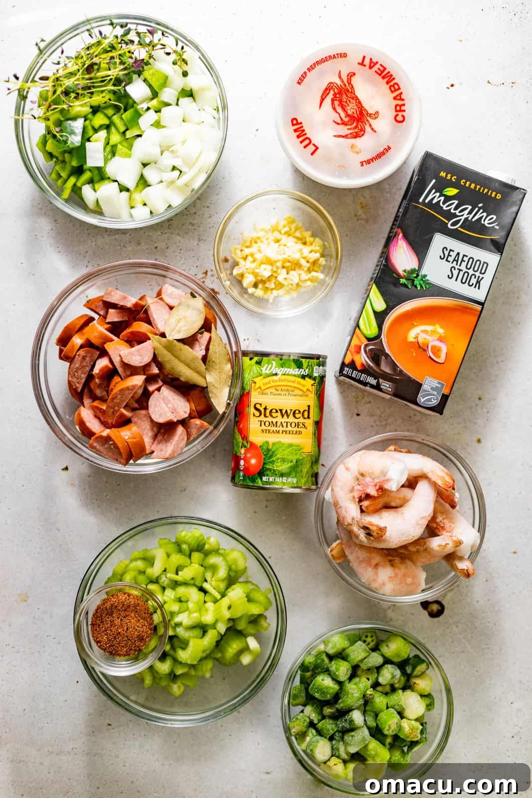 Ingredients laid out on a white surface for making seafood gumbo, including shrimp, crab meat, oils, flour, Cajun seasoning, tomatoes, celery, garlic, onion, bell pepper, seafood stock, chicken stock, bay leaves, Andouille sausage, okra, filé powder, and Worcestershire sauce.