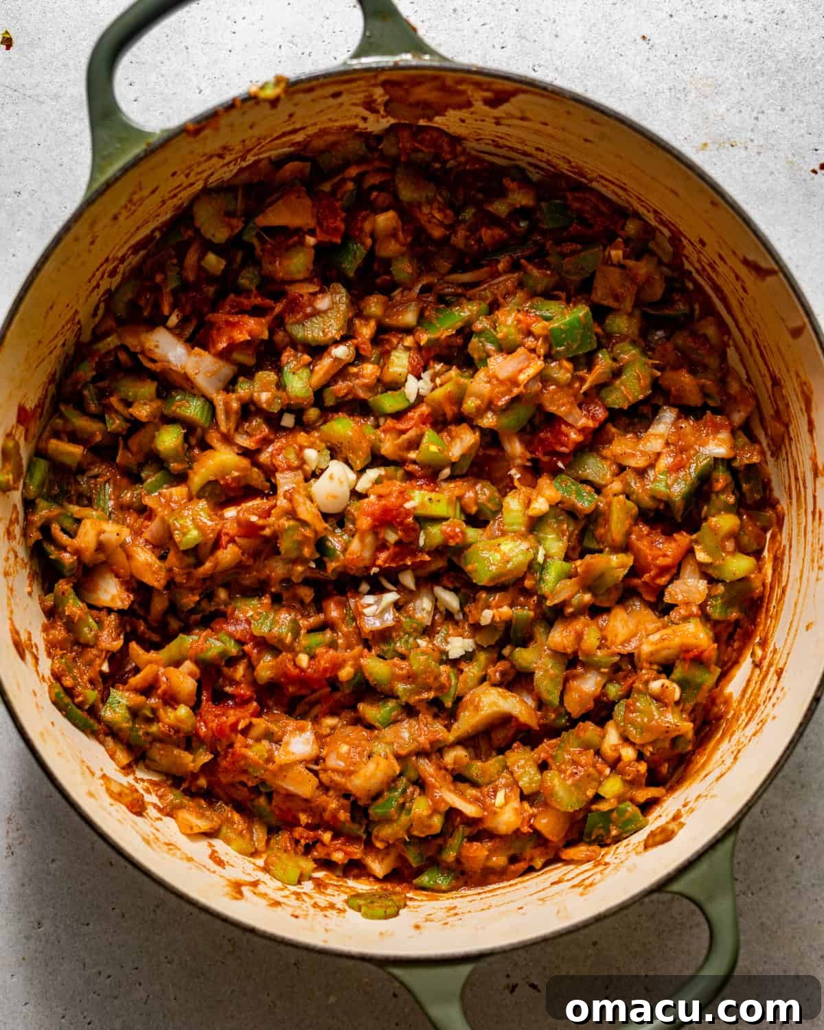 Diced bell peppers, onions, celery, garlic, stewed tomatoes, and various seasonings cooking in a dark roux within a large pot.