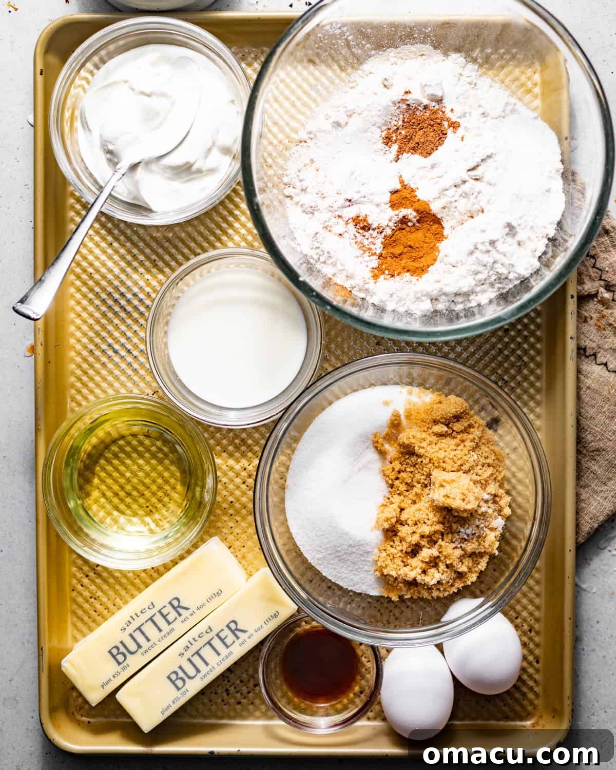Ingredients for making coffee cake muffins displayed on a baking sheet: butter, oil, eggs, milk, sour cream, vanilla extract, flour, cinnamon, nutmeg, baking powder, baking soda, and kosher salt.