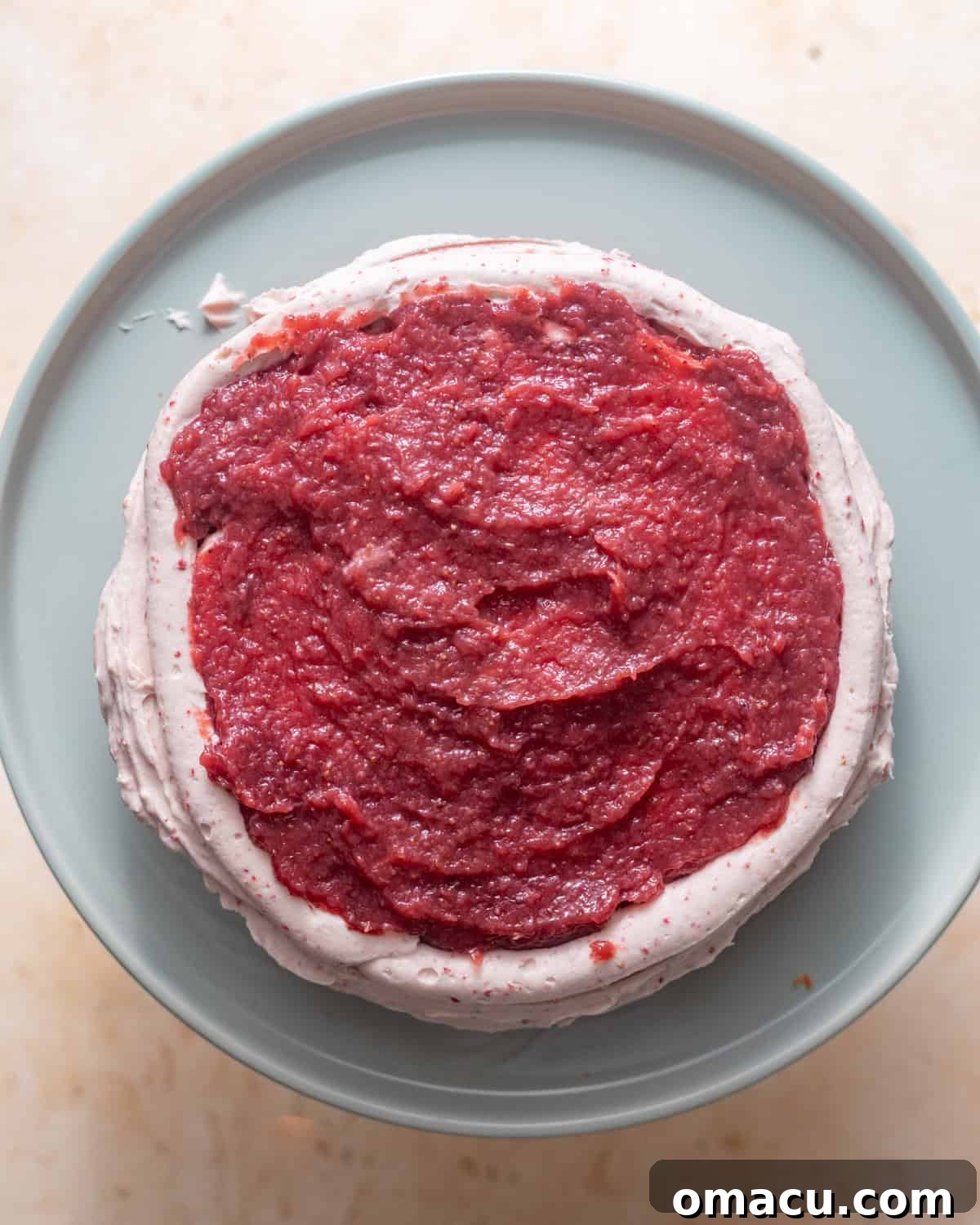 The first layer of strawberry cake on a cake stand, with a thin layer of frosting and a piped border of frosting filled with strawberry jam.
