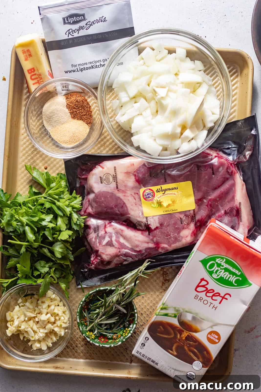 Arrangement of ingredients for beef tips and gravy on a baking sheet, including chuck roast, diced onion, minced garlic, chopped bell pepper, various seasonings, flour, beef broth, red wine, sliced celery, fresh rosemary, Sazón packet, and fresh thyme.