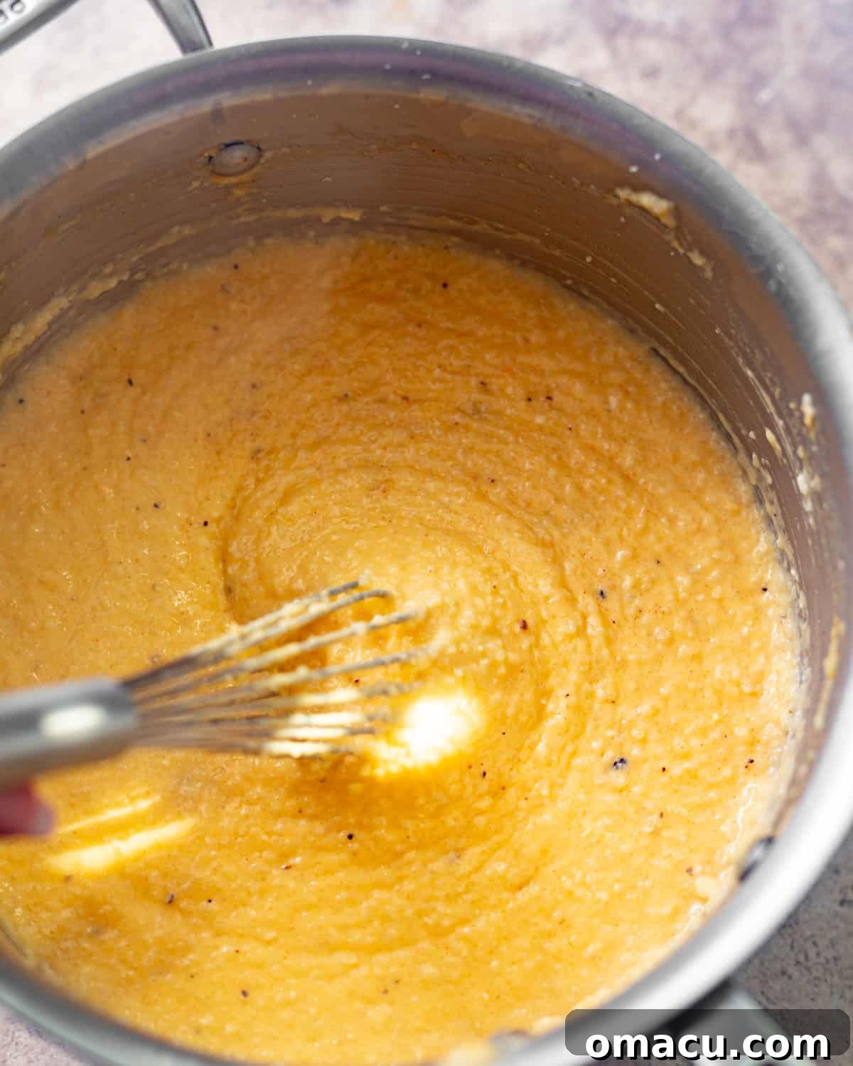 Cheesy grits in a saucepan, being whisked together with cream, cheddar cheese, and seasonings to achieve a smooth consistency.