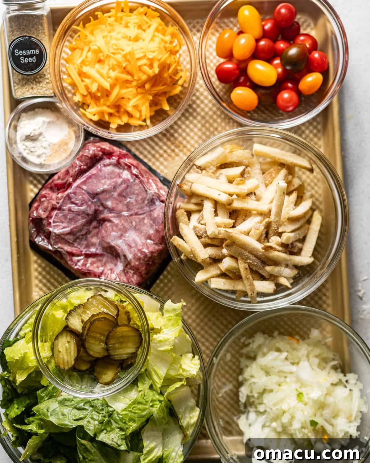 Big Mac Salad ingredients spread out on a baking sheet, including ground beef, diced onion, seasonings, pickles, cherry tomatoes, fresh lettuce, french fries, shredded cheese, and a sprinkle of sesame seeds.
