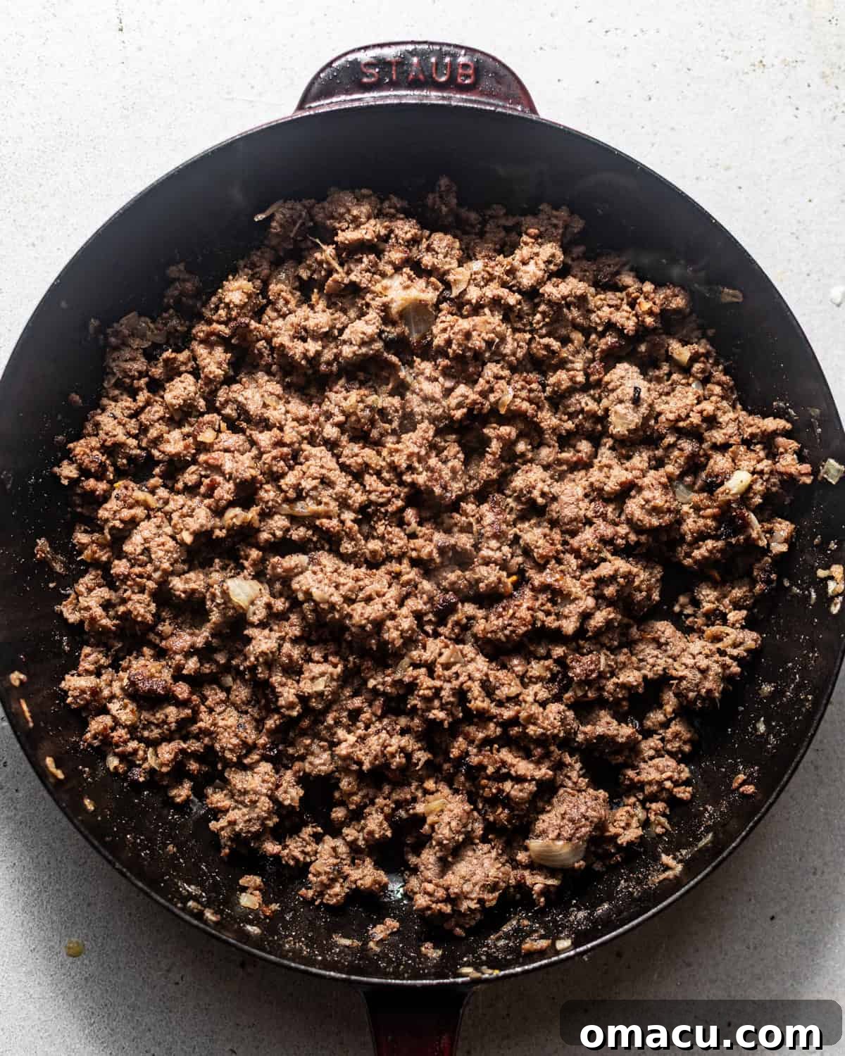 Ground beef cooking in a large skillet with grated onion, seasonings, yellow mustard, and Worcestershire sauce, being stirred to combine.