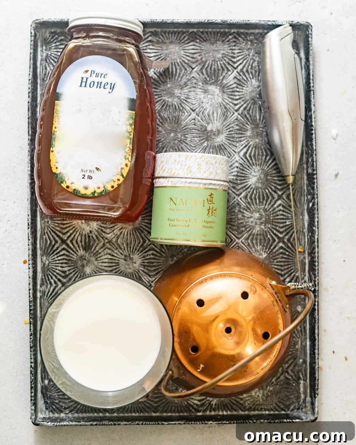 Ingredients for making an iced matcha latte arranged on a serving board: honey, ceremonial grade matcha powder, and a glass of milk.