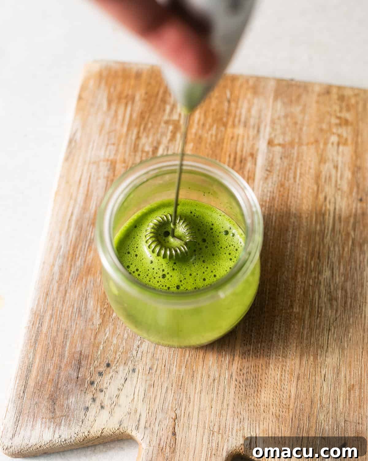 A handheld milk frother mixing warm water, matcha powder, and honey in a small cup, creating a frothy green paste.