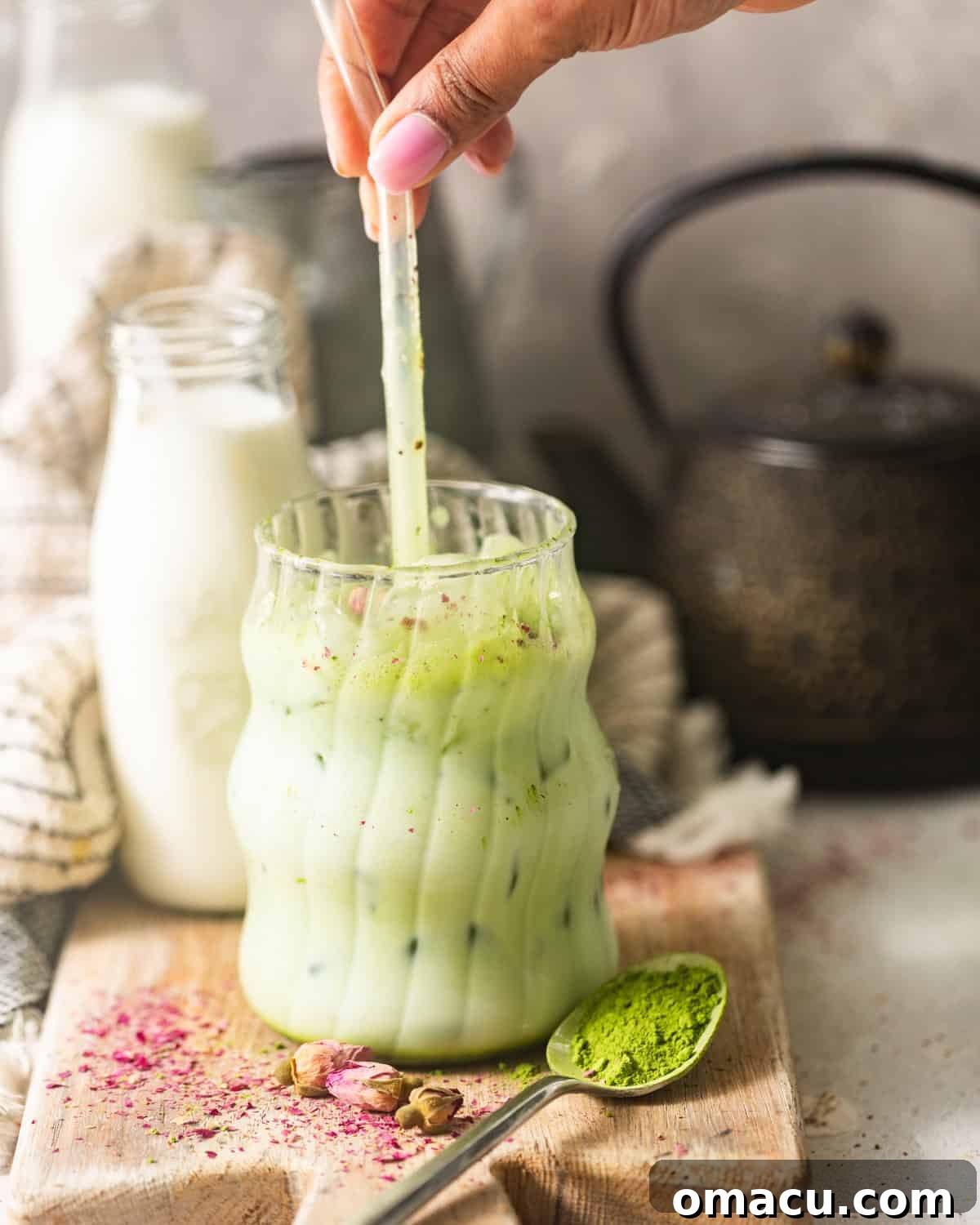 A hand pouring the whisked matcha mixture into a tall glass filled with ice and milk, ready to be stirred and enjoyed.