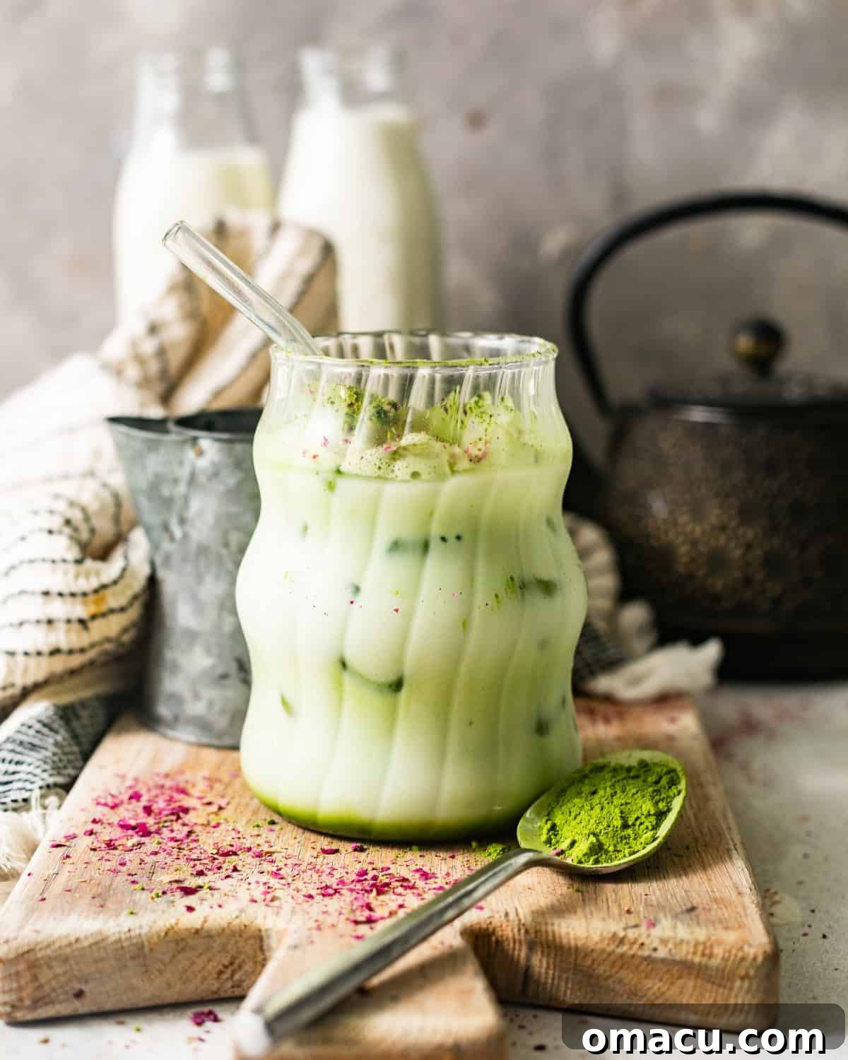 A pristine Iced Matcha Latte in a glass with a straw, sitting on a wooden serving board alongside small bottles of milk, emphasizing its homemade appeal.