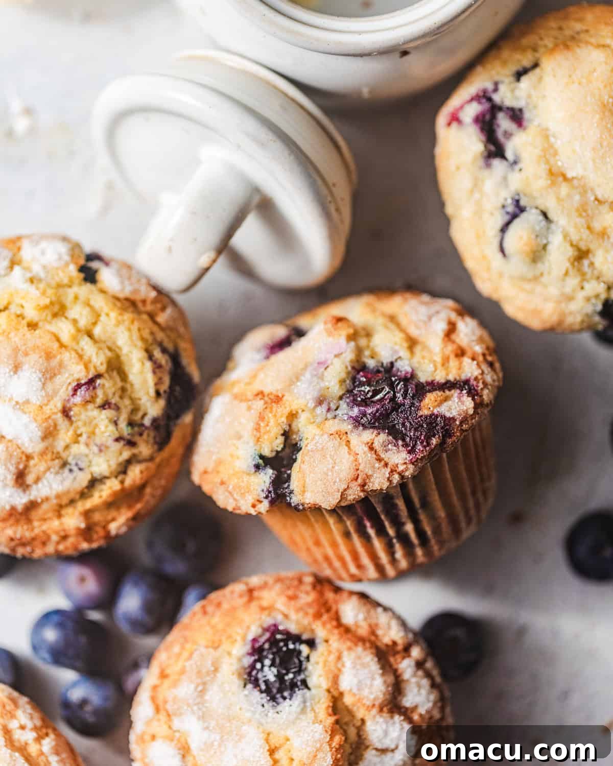 Freshly baked bakery-style blueberry muffins on a white surface with fresh blueberries scattered around. The muffins have golden, domed tops and a perfect crumb.