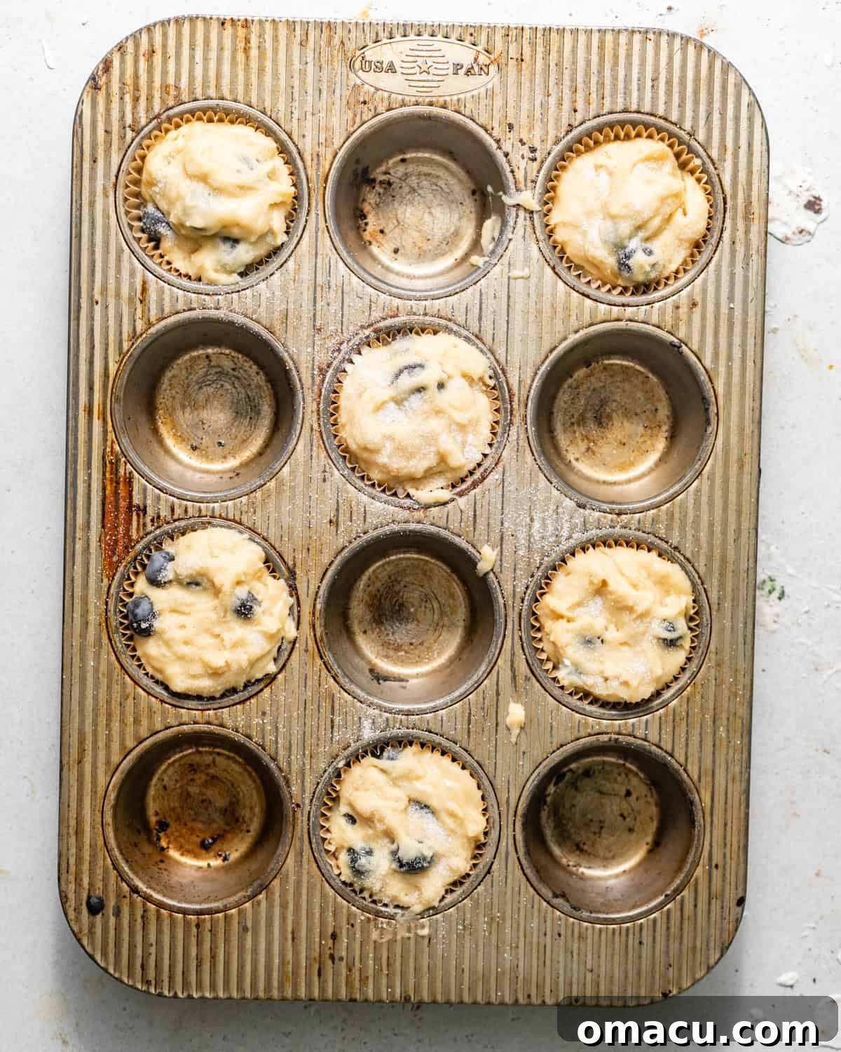 Muffin batter scooped into a muffin pan, filling the cups to the top, with extra blueberries and sugar sprinkled on top, ready for baking.