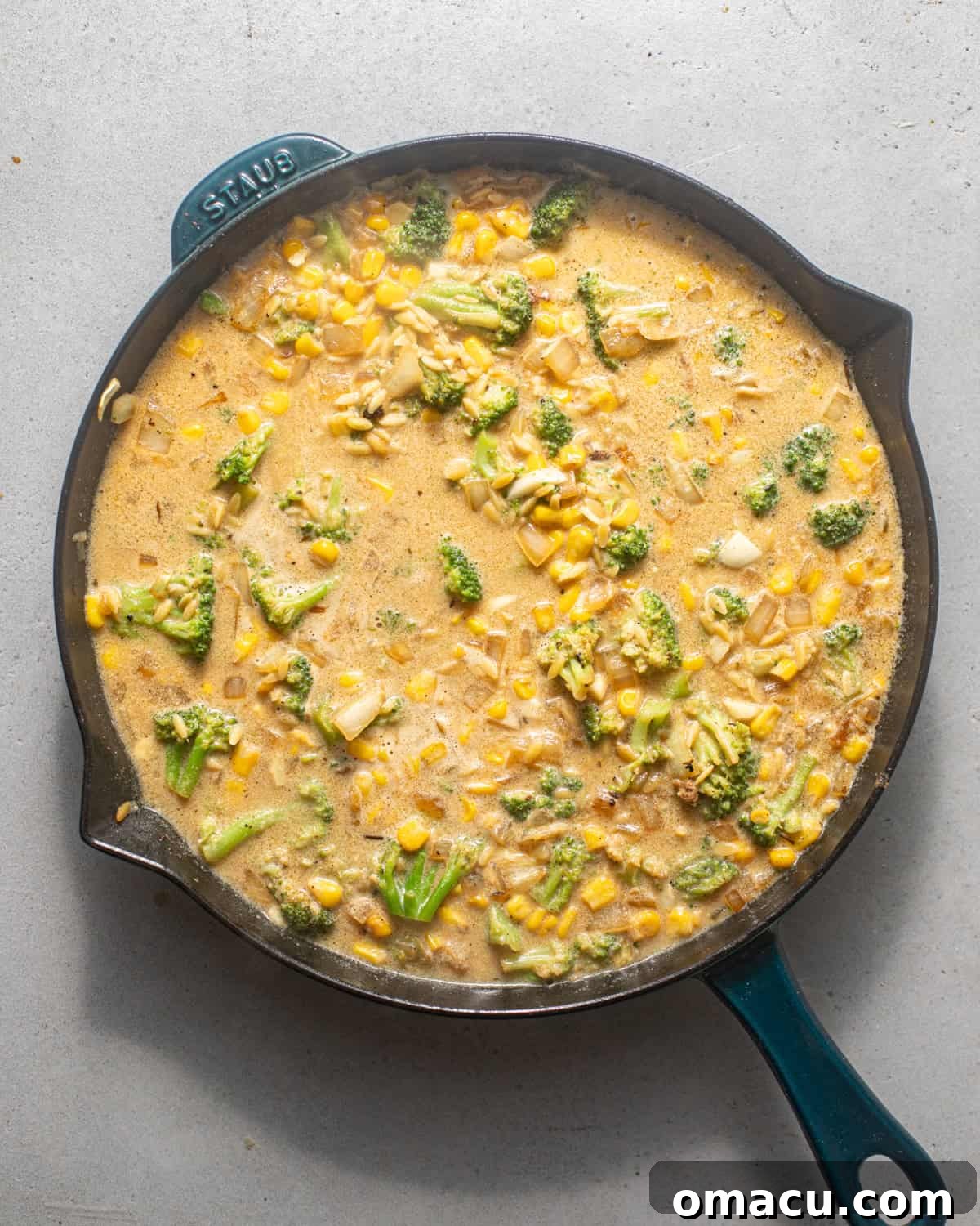 Chicken broth being poured into a skillet containing sautéed vegetables, onions, garlic, and toasted orzo.