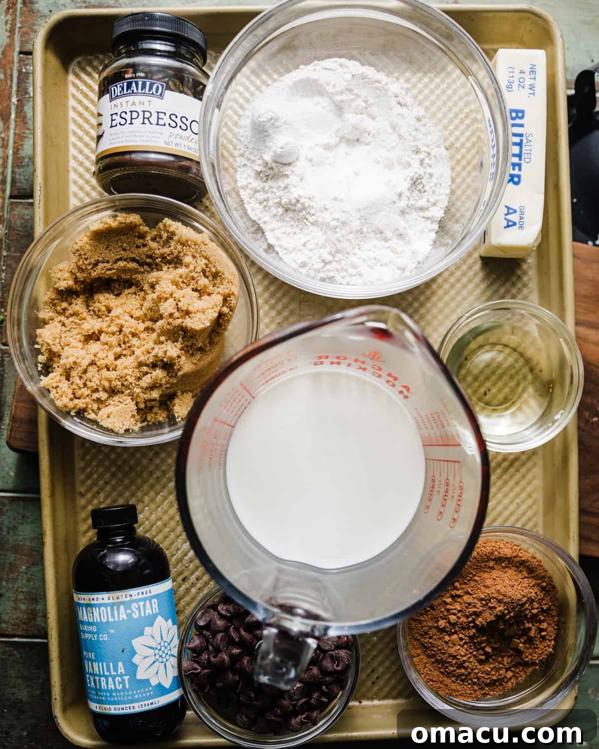 Essential ingredients laid out for making a rich chocolate donut cake, including flour, cocoa powder, sugars, vanilla, oils, butter, eggs, espresso powder, powdered sugar, milk, and salt.