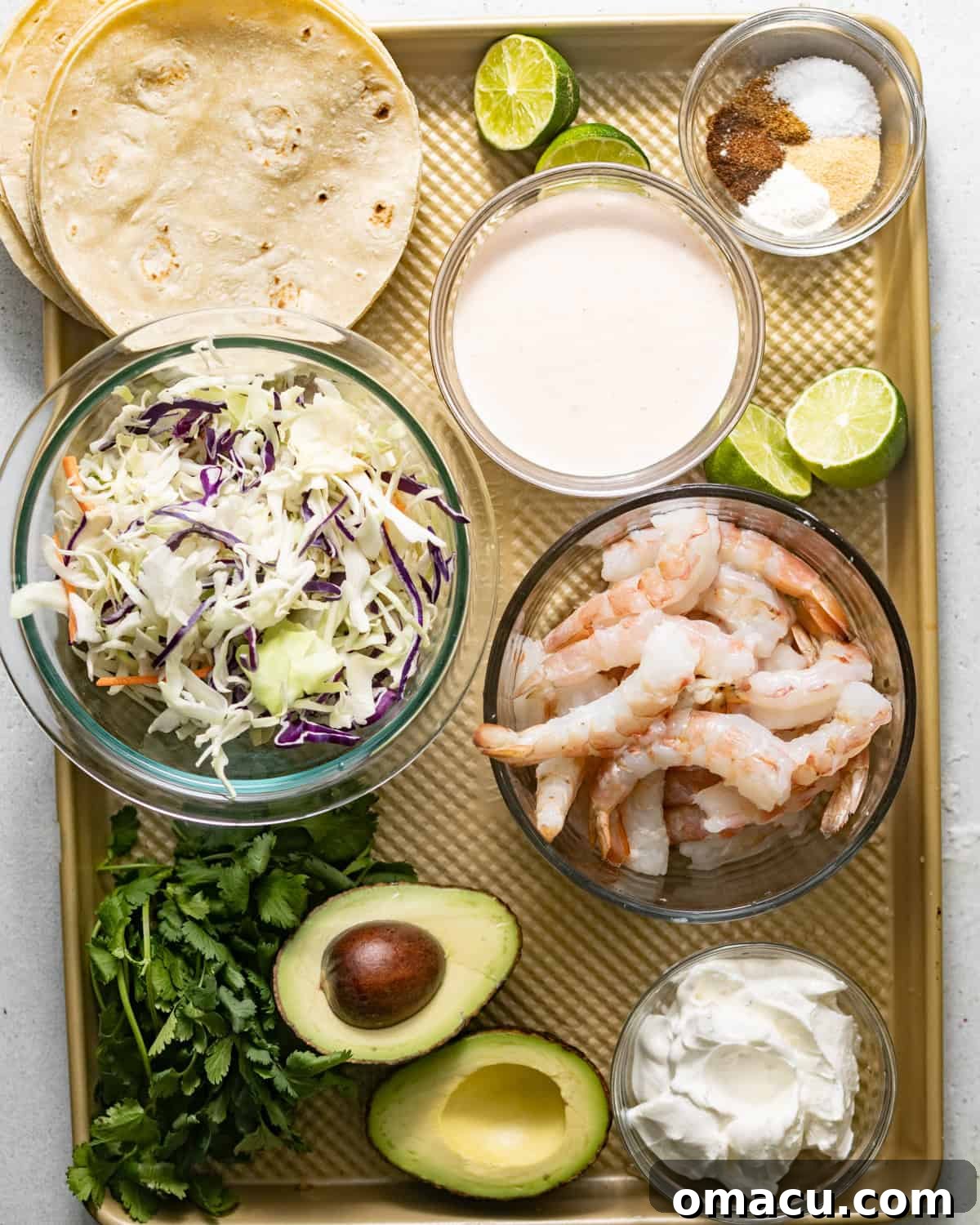 Ingredients laid out on a baking sheet to make baja shrimp tacos, including fresh shrimp, seasonings, coleslaw mix, ripe avocado, cilantro, limes, tortillas, sour cream, and hot sauce.