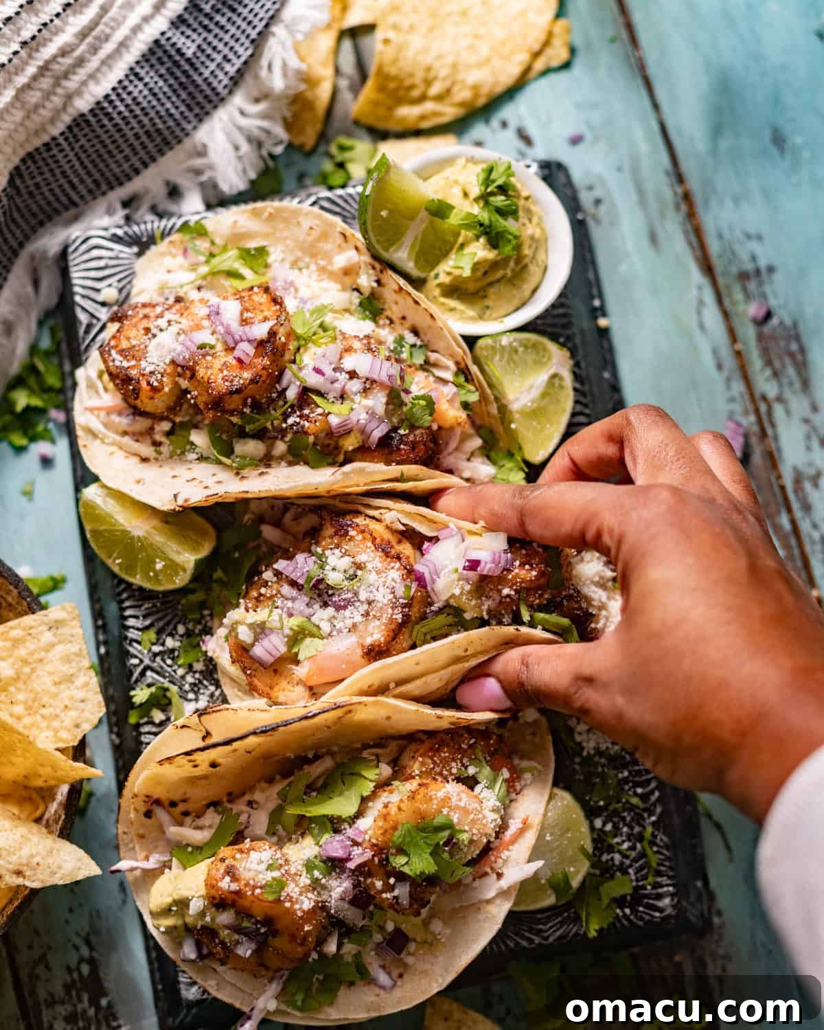 A hand reaching for a fully assembled Baja shrimp taco, showcasing the layers of shrimp, slaw, and avocado crema.