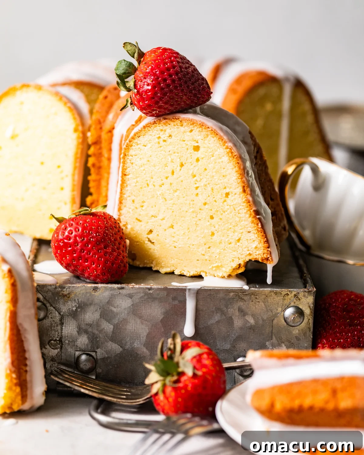 A perfectly sliced piece of whipping cream pound cake on a white plate, garnished with fresh strawberries, sitting on a rustic wooden table with additional plates and berries in the background.