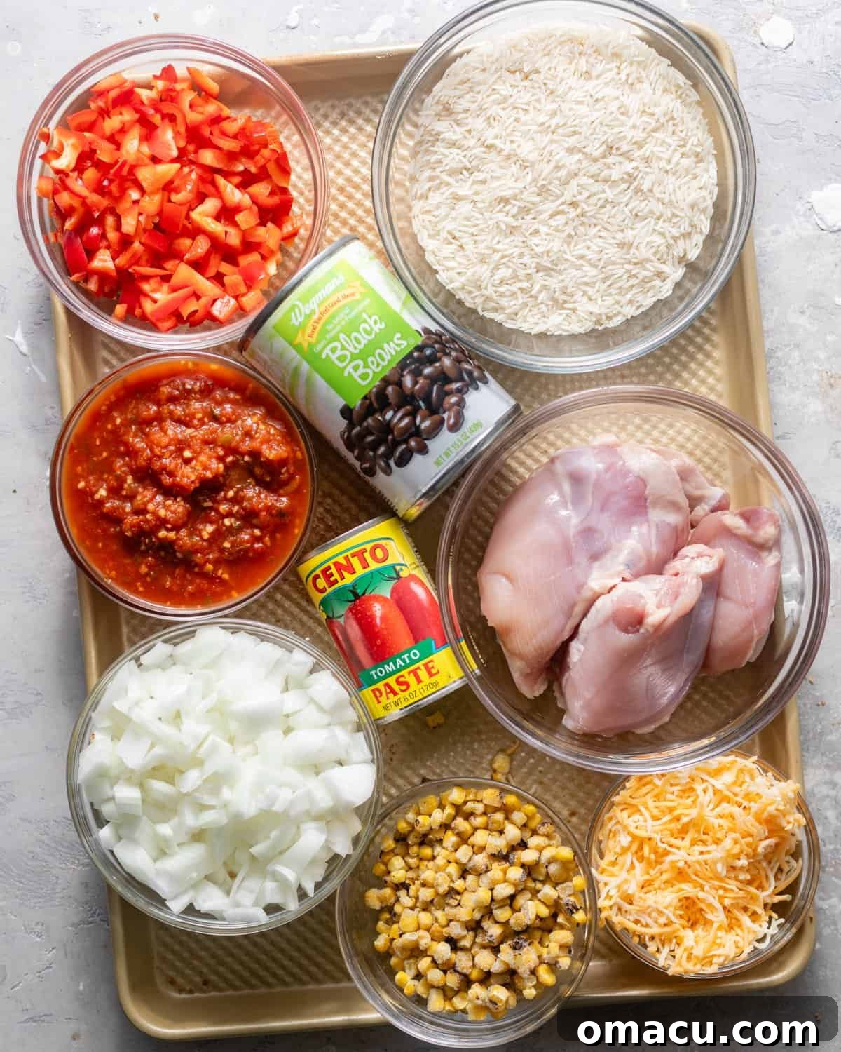 A vibrant flat lay of ingredients laid out on a tray for Mexican chicken and rice, including uncooked rice, canned black beans, chopped bell pepper, salsa, onions, garlic, frozen corn, boneless skinless chicken thighs, tomato paste, various seasonings, tortilla chips, Modelo beer, and chicken broth.