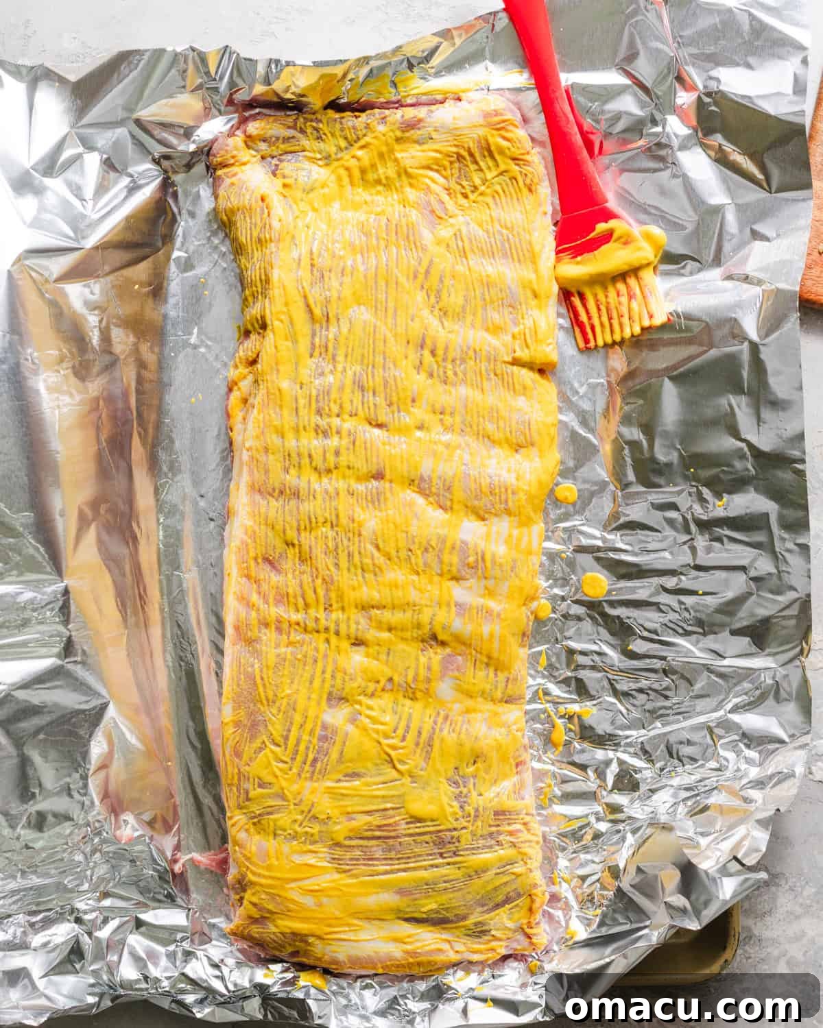 Pork ribs on parchment paper being brushed with yellow mustard.