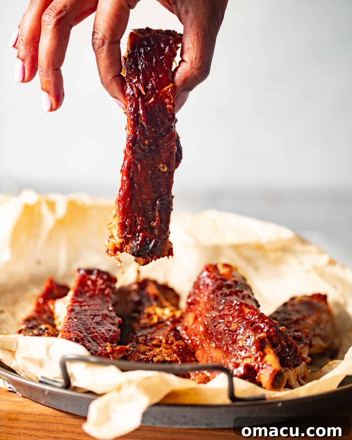 A hand holding a single perfectly cooked rib above a plate piled with more ribs.