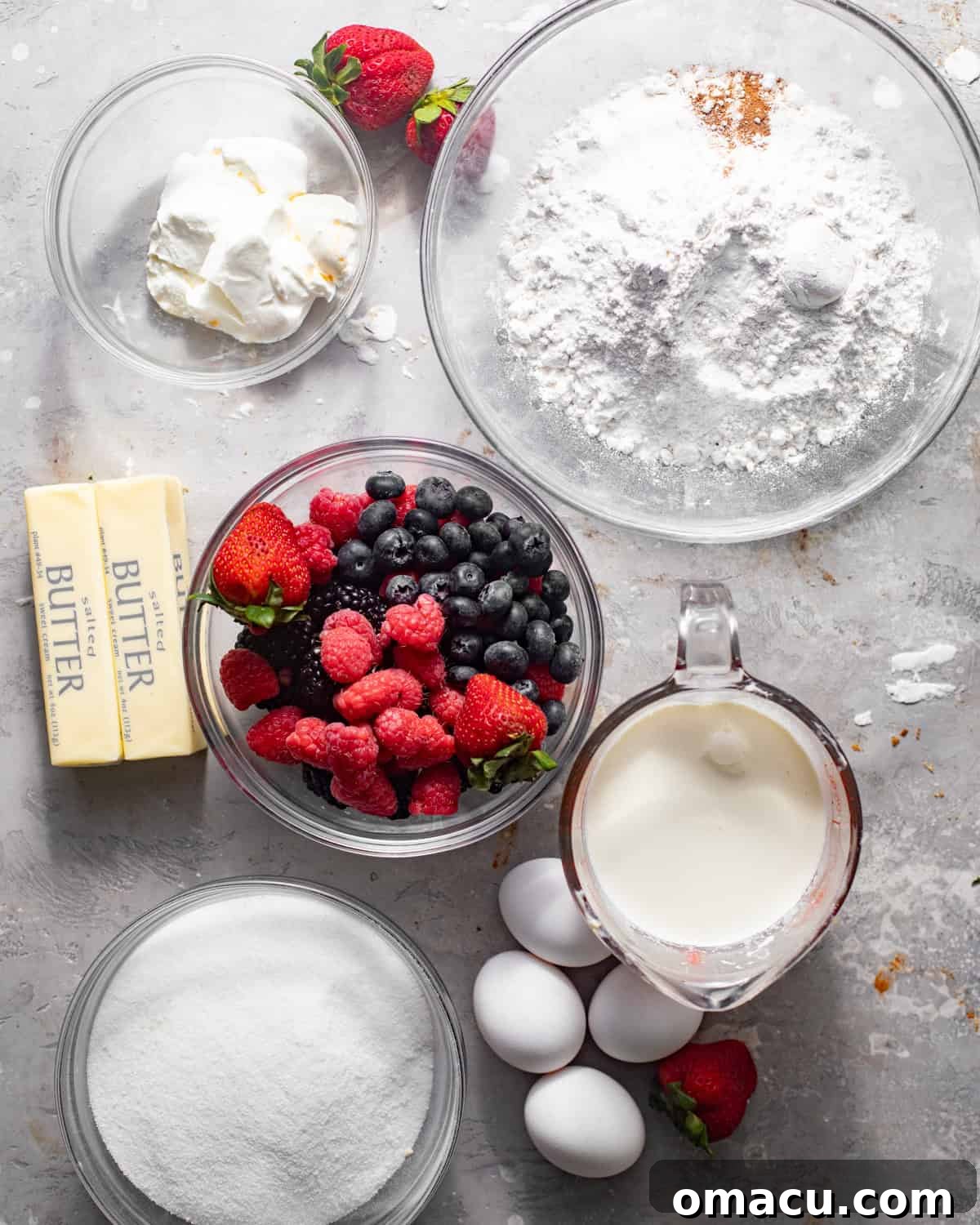 A curated selection of fresh ingredients laid out on a clean white surface, showcasing heavy cream, various berries, flour, butter, and more, all ready for making the Berry Chantilly Cake.