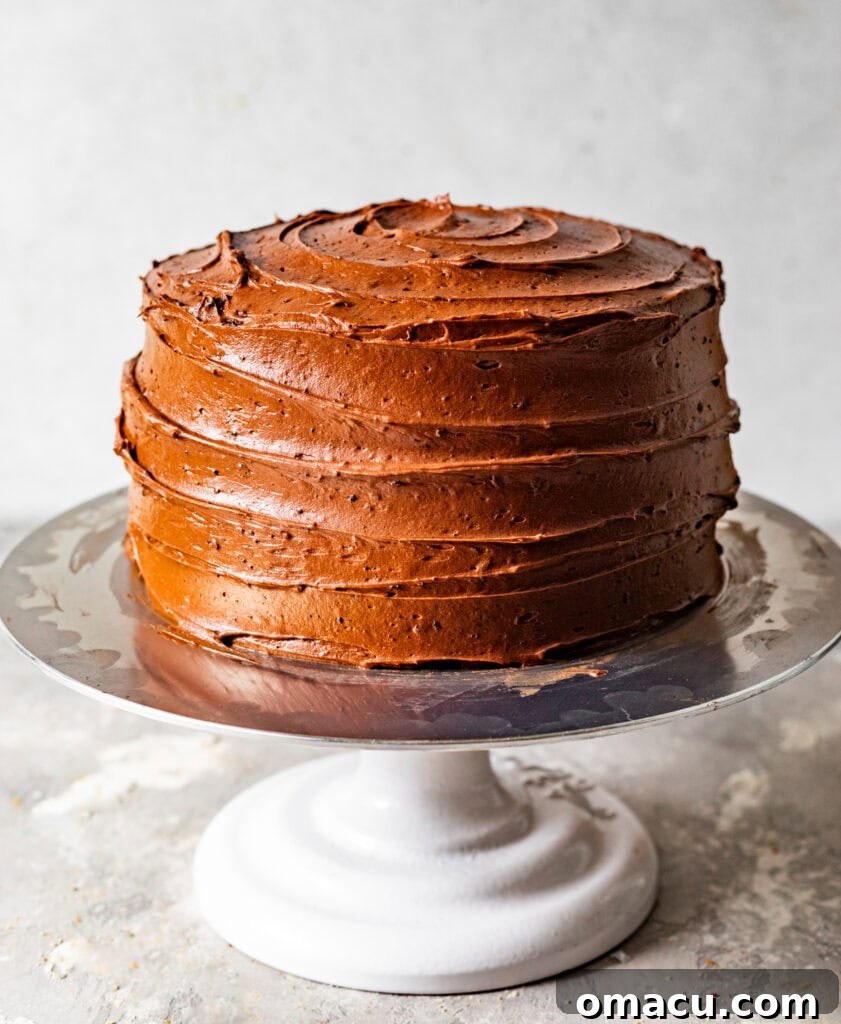 A three-layer chocolate cake is being assembled on a cake stand, with even layers of chocolate cream cheese frosting spread between each cake layer, and a thin crumb coat applied to the outside.