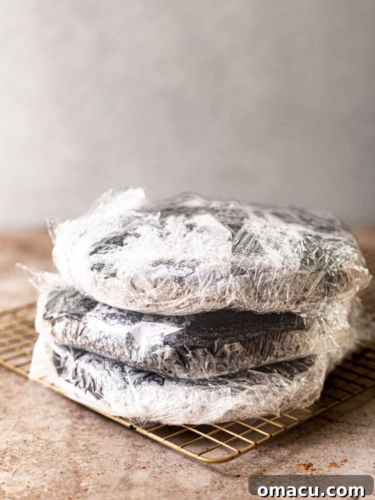 Three cooled chocolate cake layers are individually wrapped in plastic wrap on a wire rack, preserving their moisture before frosting.