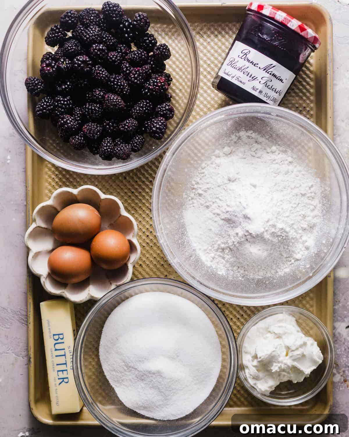 Ingredients laid out on a baking tray for blackberry coffee cake: butter, sugar, eggs, fresh blackberries, flour, salt, vanilla extract, baking powder, and blackberry preserves.