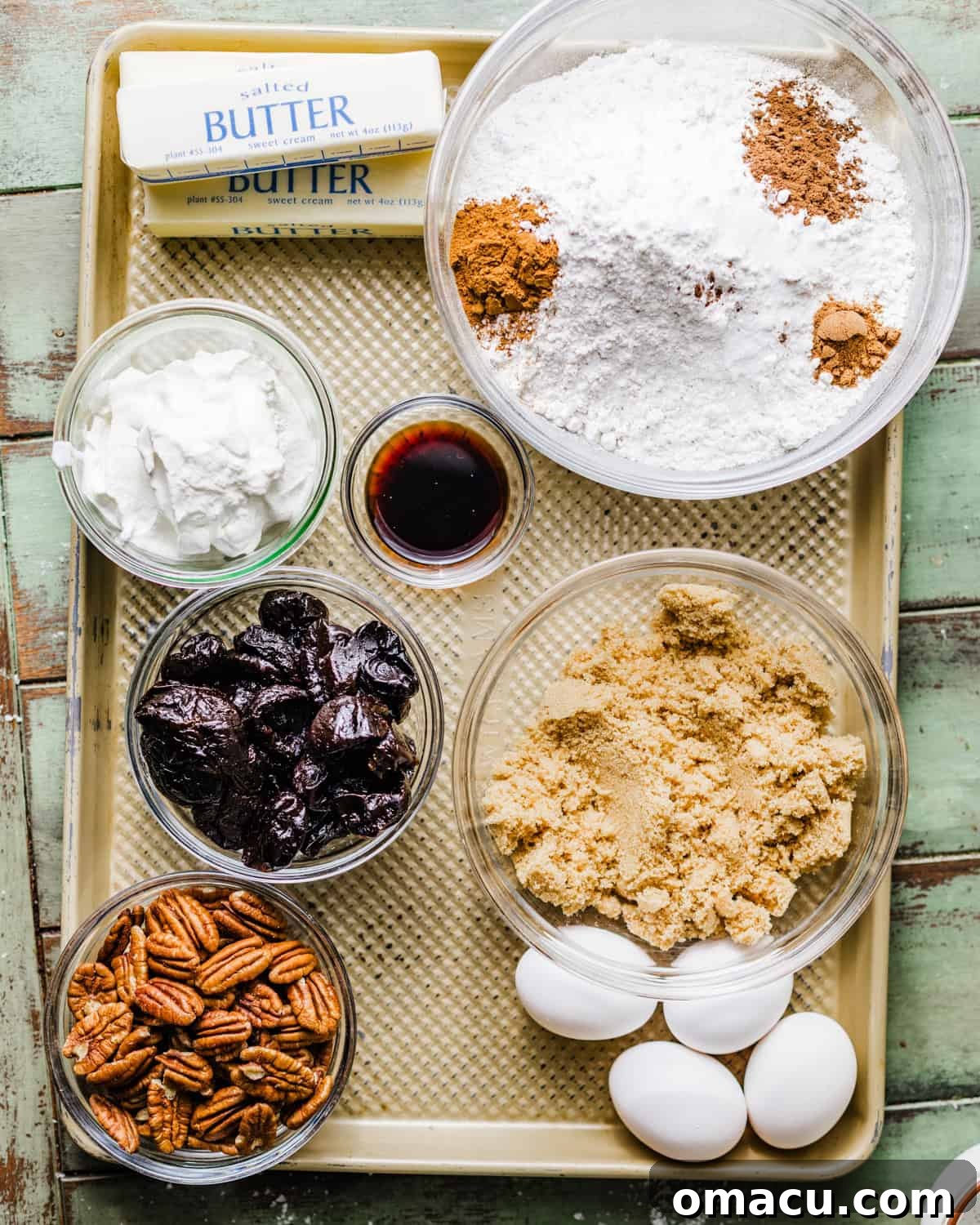 Ingredients laid out on a baking sheet for prune cake: all purpose flour, brown sugar, granulated sugar, eggs, pecans, prunes, vanilla extract, shortening, butter, allspice, cinnamon, nutmeg, salt, whole milk, and sour cream.