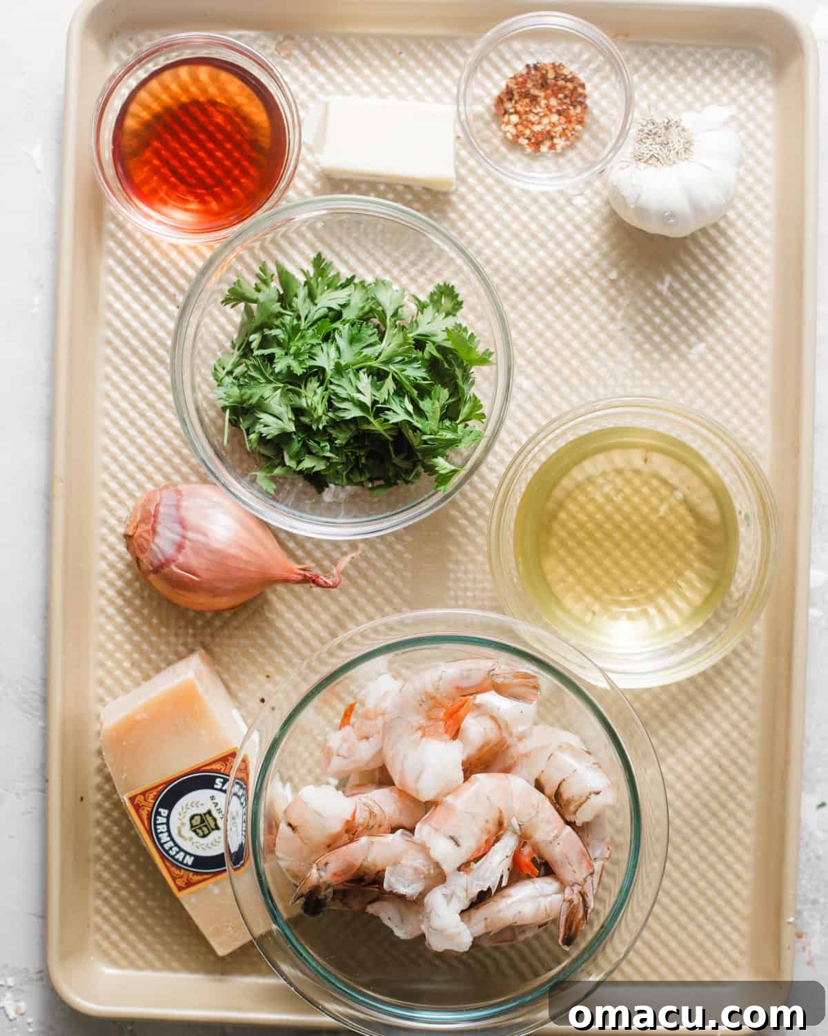 A collection of fresh ingredients neatly arranged on a baking sheet, including jumbo shrimp, vibrant parsley, a bowl of seasoning, parmesan cheese, olive oil, onion, shallot, red wine vinegar, and lemon juice – all set to make shrimp chimichurri.