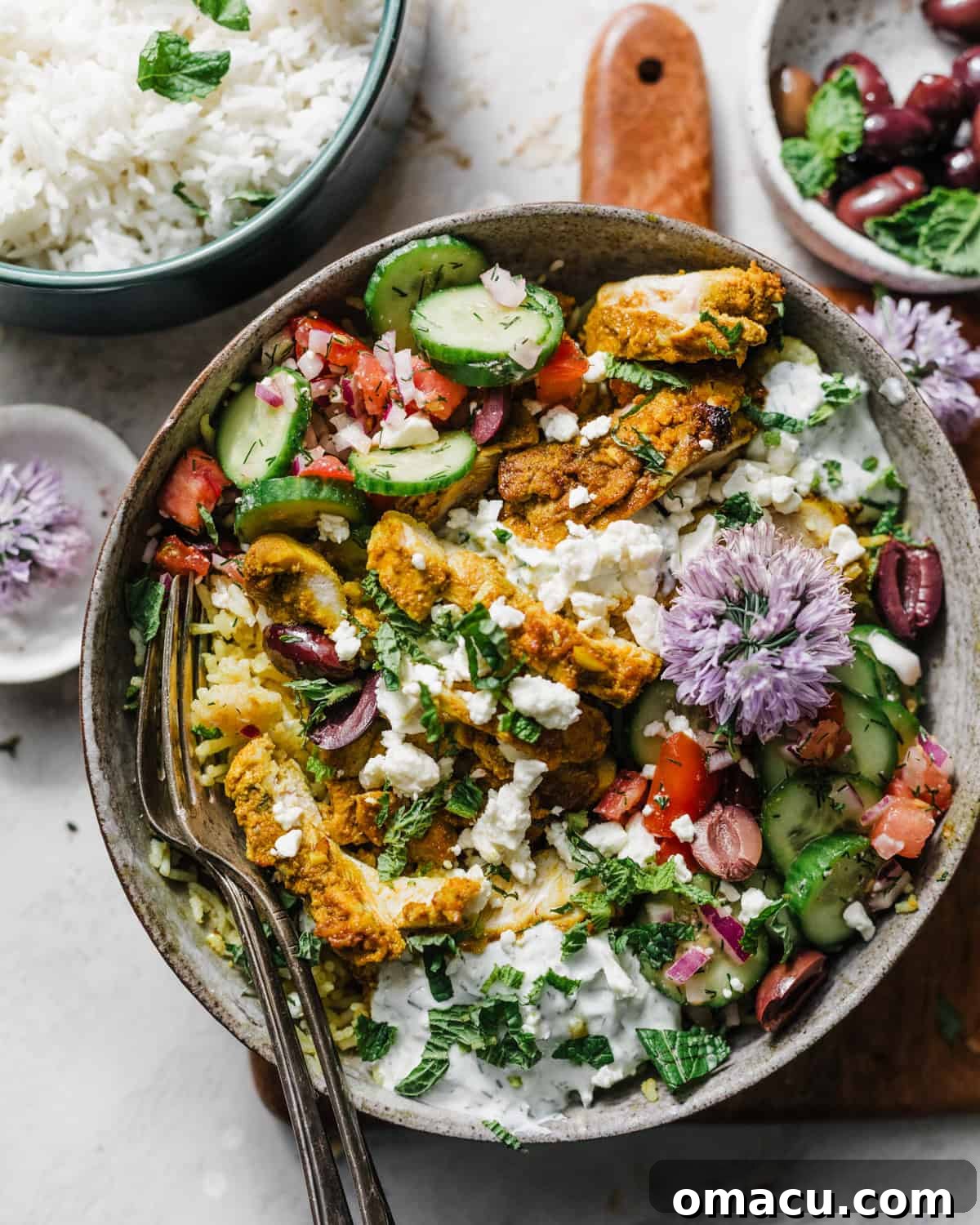 A vibrant Mediterranean Chicken Bowl on a serving board, accompanied by a side bowl of fluffy rice and sliced Kalamata olives, showcasing the fresh and colorful ingredients.
