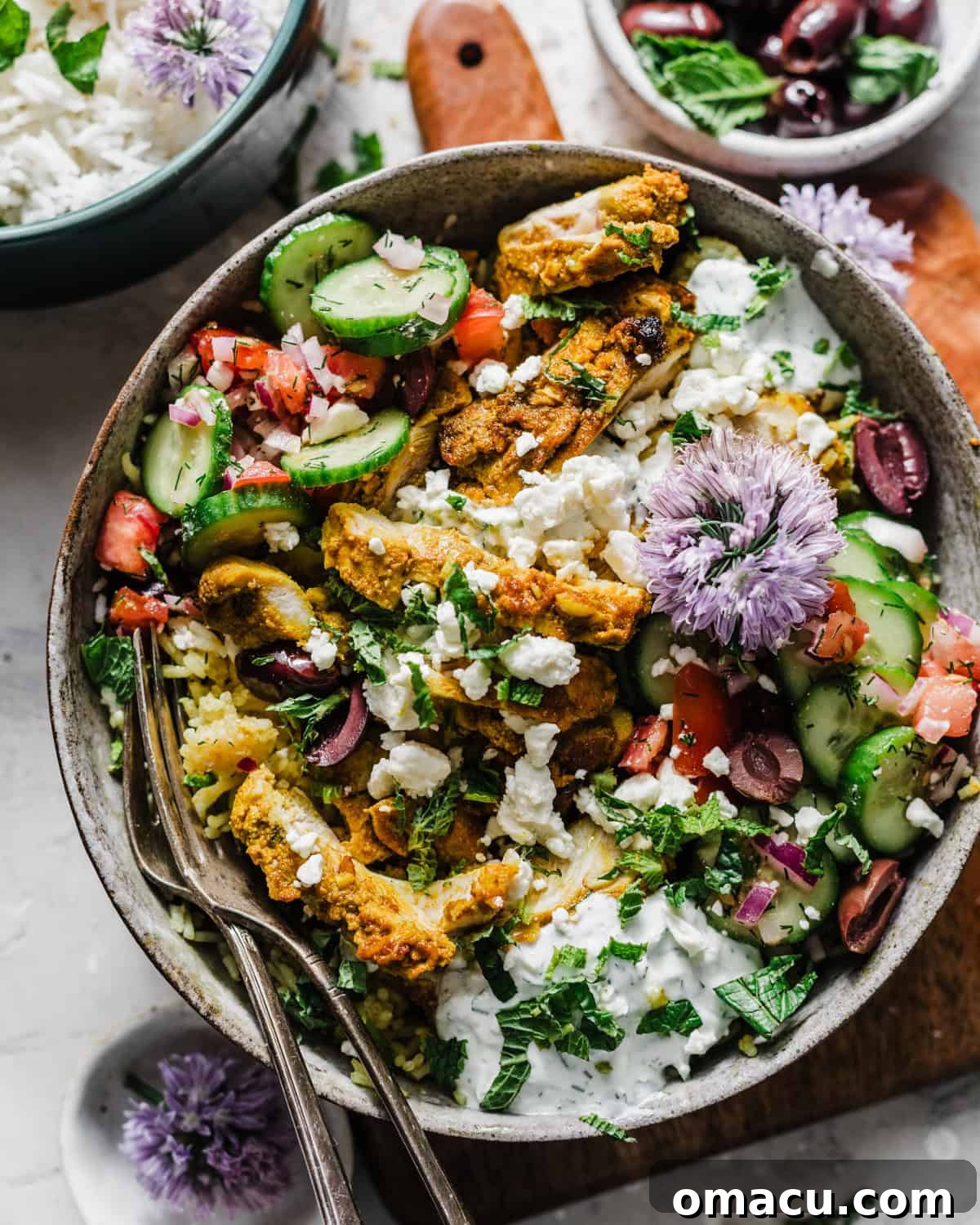 Overhead photo of a beautifully arranged Mediterranean Chicken Bowl on a serving board, surrounded by forks, crumbled feta cheese, Kalamata olives, and fresh cucumber salad, highlighting its freshness and appeal.