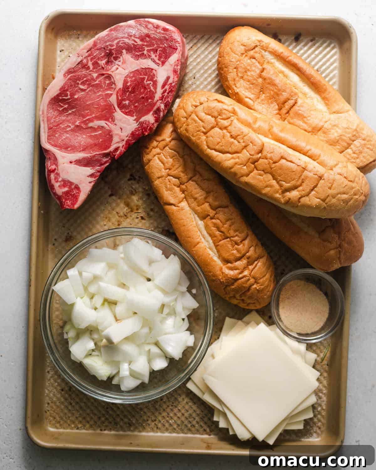 A visual display of all necessary ingredients for making a classic Philly cheesesteak, including ribeye steak, onions, American cheese, a selection of seasonings, and an authentic hoagie roll, arranged neatly on a baking sheet.