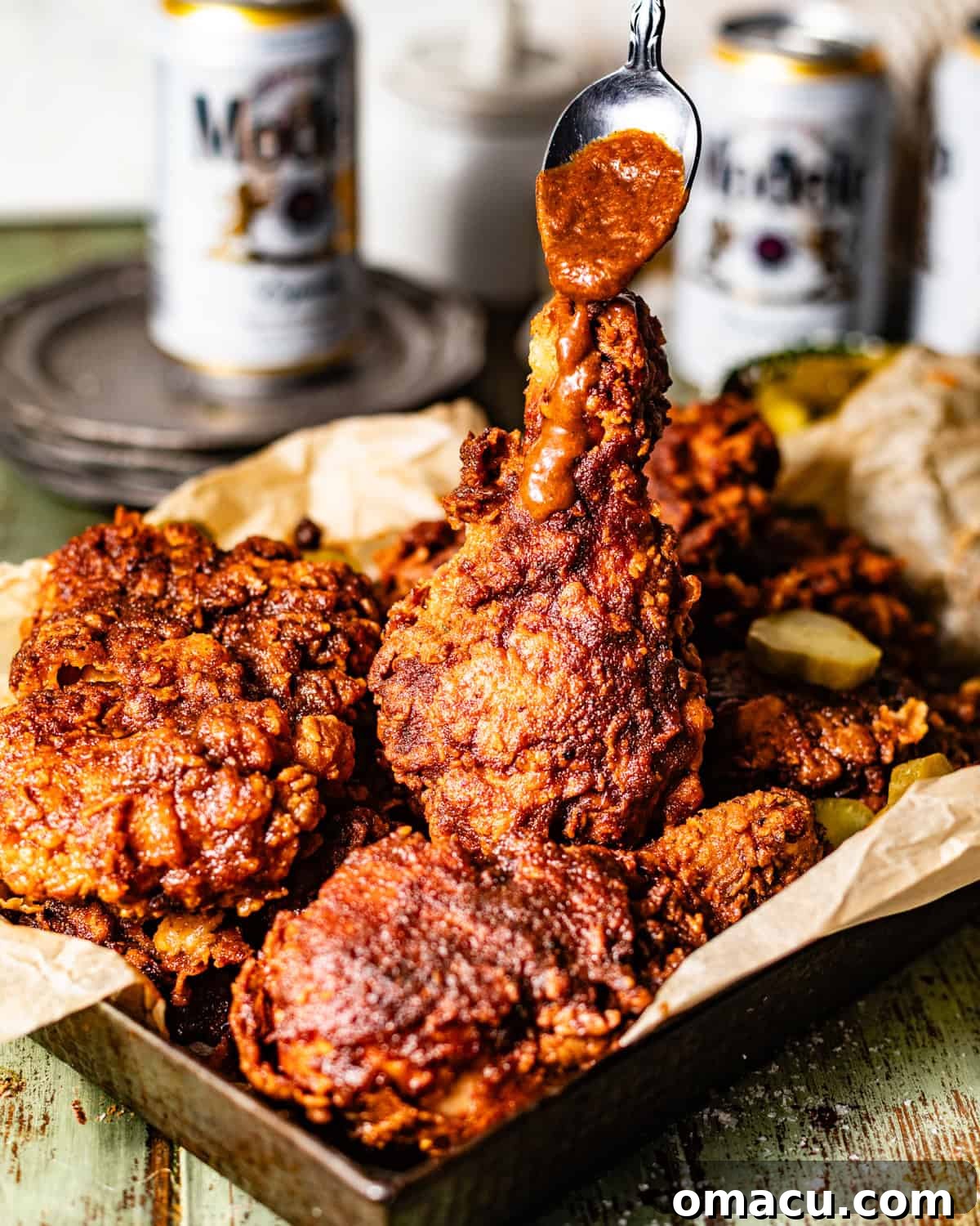 A serving basket filled with crispy Nashville Fried Chicken, with a spoon generously drizzling the spicy, glossy sauce over the top, ready to be devoured.