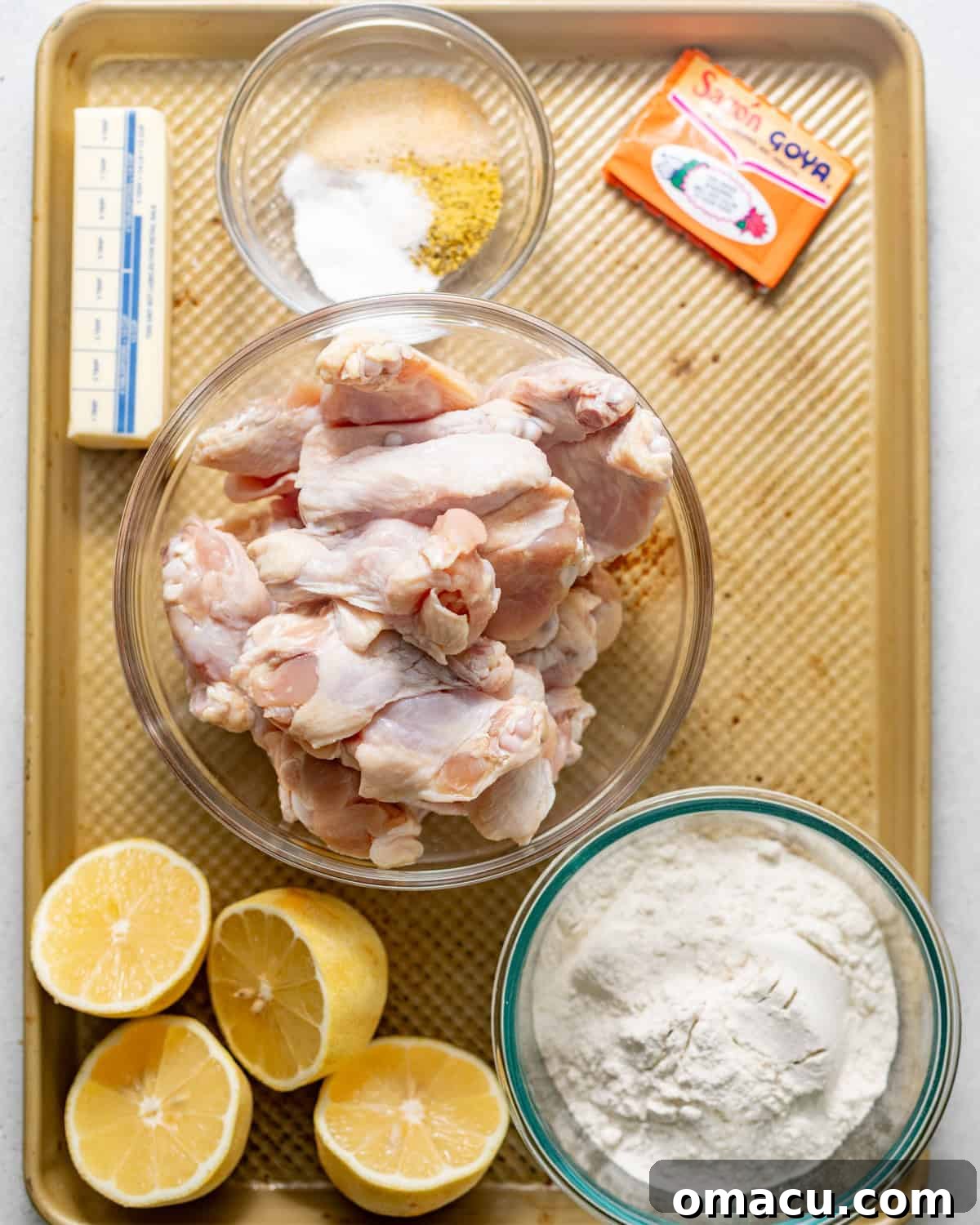 A vibrant display of key ingredients for lemon pepper wings laid out on a baking sheet, including raw chicken wings, sazon seasoning, fresh lemon juice and zest, all-purpose flour, lemon pepper seasoning, garlic and onion powder, mustard, hot sauce, butter, and honey.