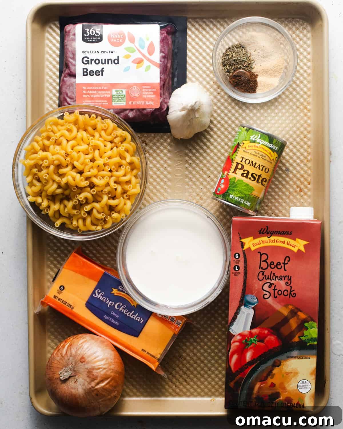 All the key ingredients for homemade Hamburger Helper laid out on a baking sheet: ground beef, elbow macaroni, milk, cheddar cheese, onion, garlic, various seasonings, beef broth, BBQ sauce, Worcestershire sauce, and tomato paste.