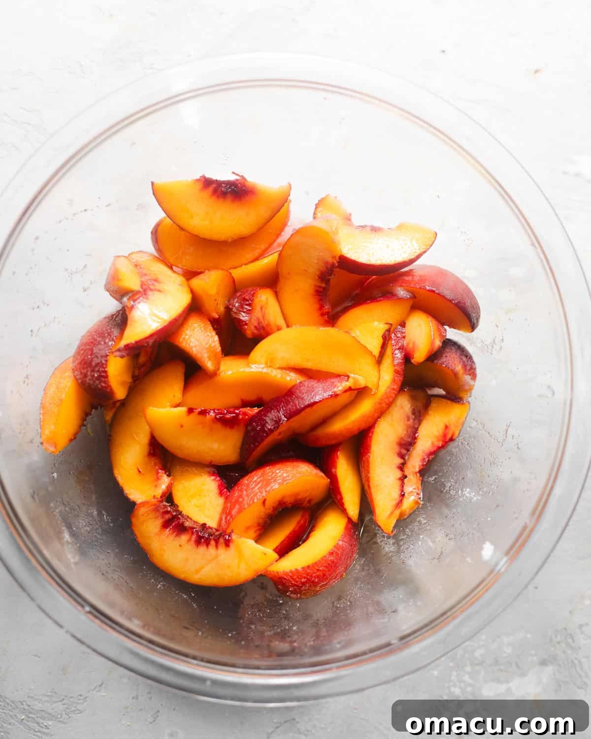 Sliced peaches macerating in a glass bowl, glistening with brown sugar and lemon juice.