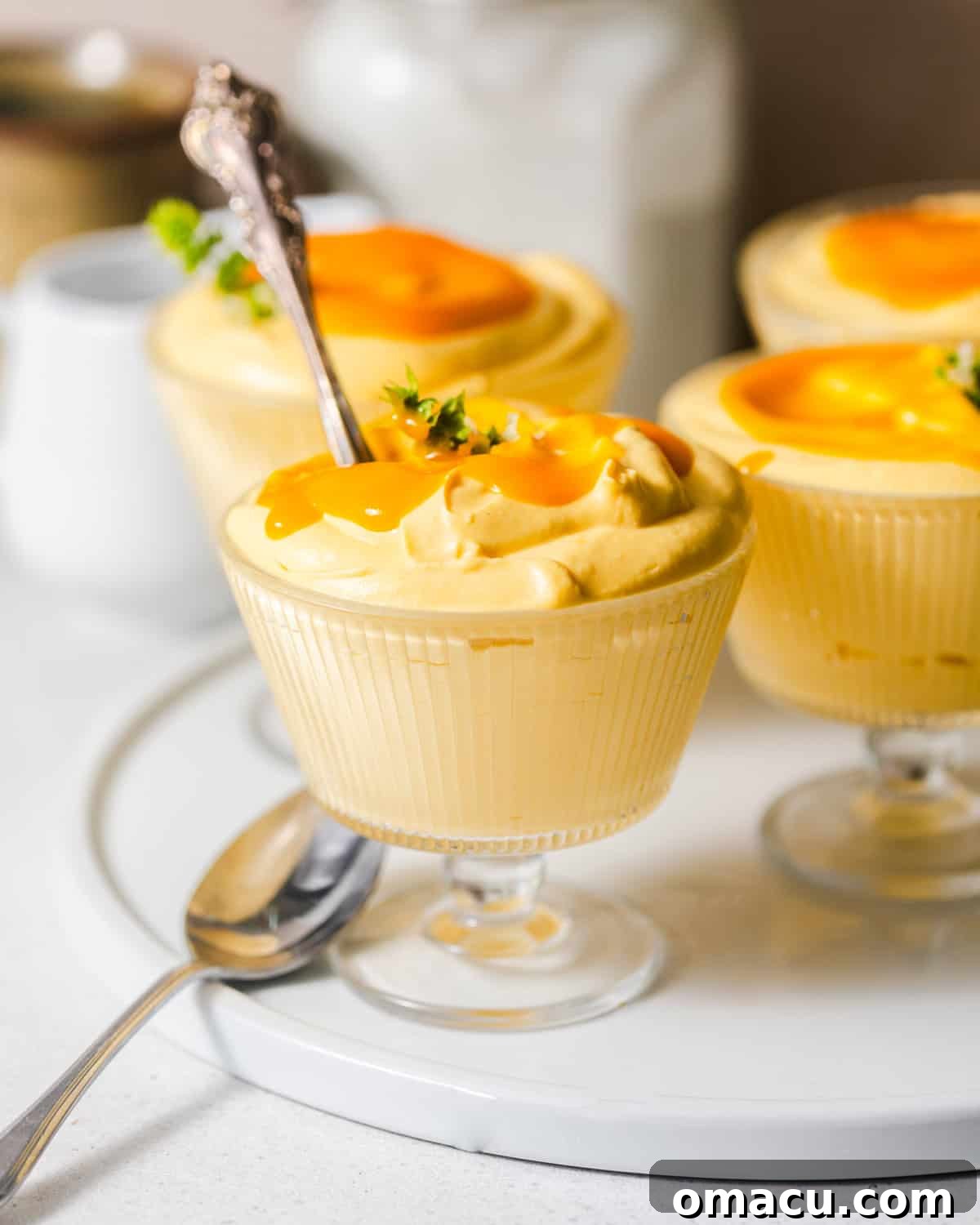 Individual servings of creamy mango mousse in clear glass cups, garnished and ready to be enjoyed on a serving plate with spoons.