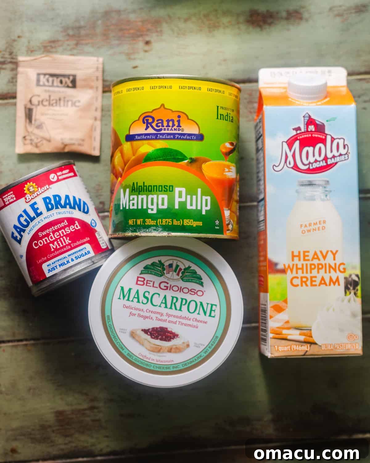 A curated selection of fresh ingredients laid out on a clean surface, including a can of Alphonso mango puree, a tub of mascarpone cheese, a can of sweetened condensed milk, a packet of gelatin, fresh lemons, and a carton of heavy cream, ready for making mango mousse.