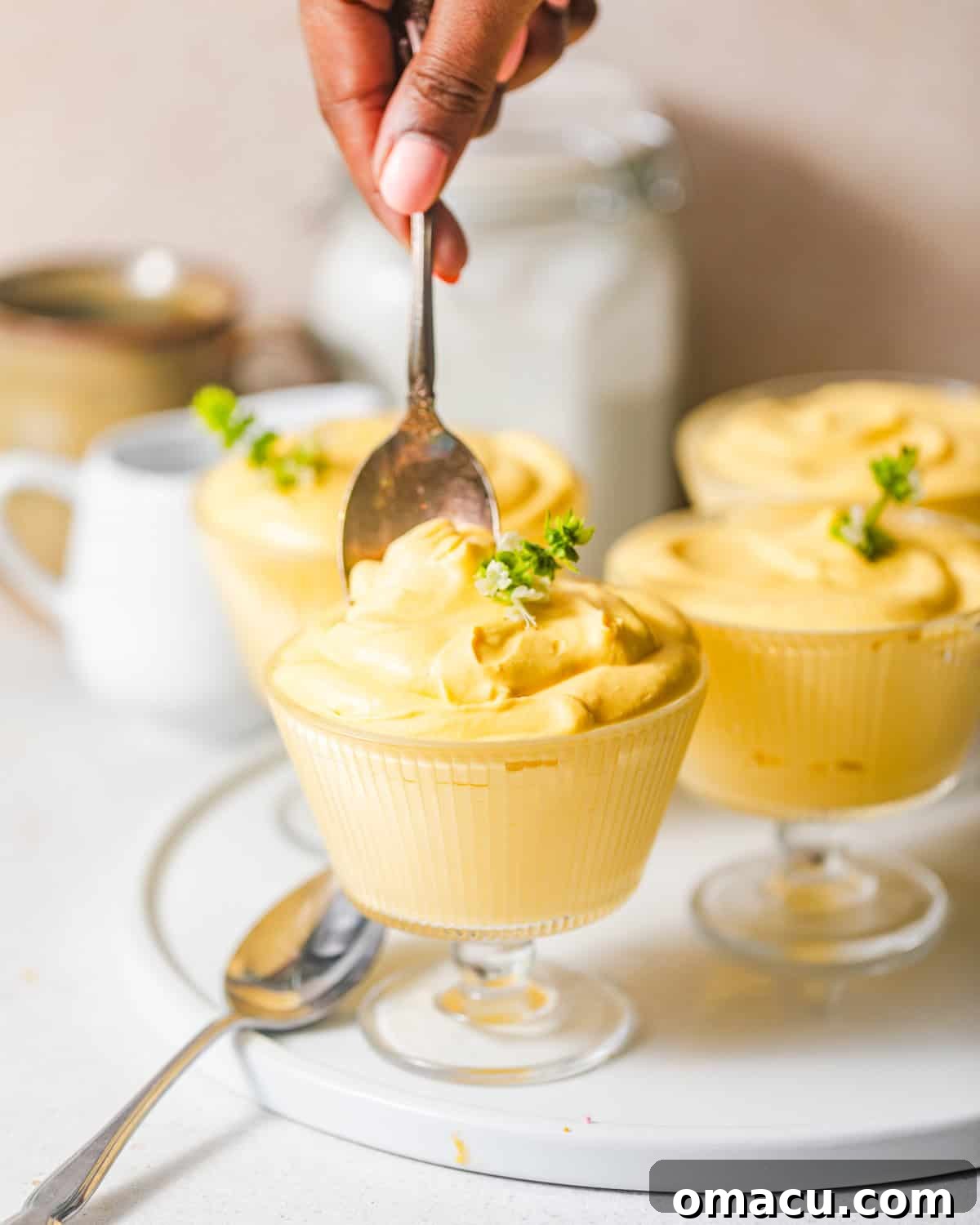 A hand carefully spooning the fluffy mango mousse into individual glass serving cups, preparing them for chilling.