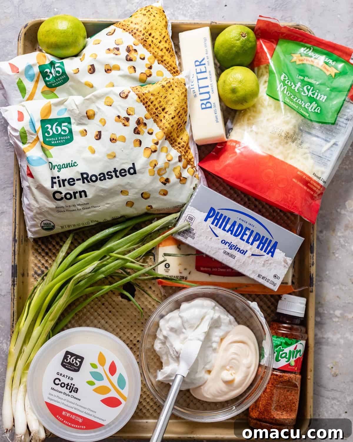 All the fresh ingredients laid out on a baking tray, including corn, limes, garlic, scallions, and various cheeses, ready to be prepared for the street corn dip.