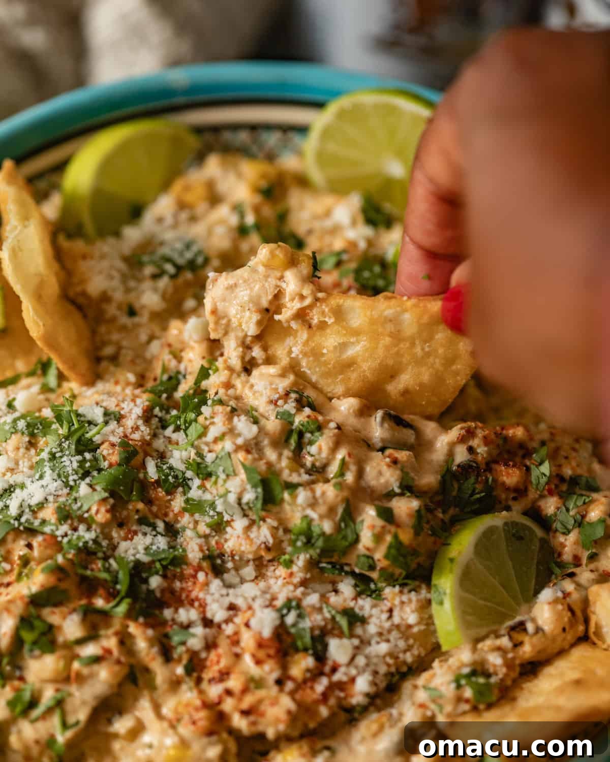 A hand holding a tortilla chip, dipping it into the creamy, cheesy Mexican street corn dip in a bowl.