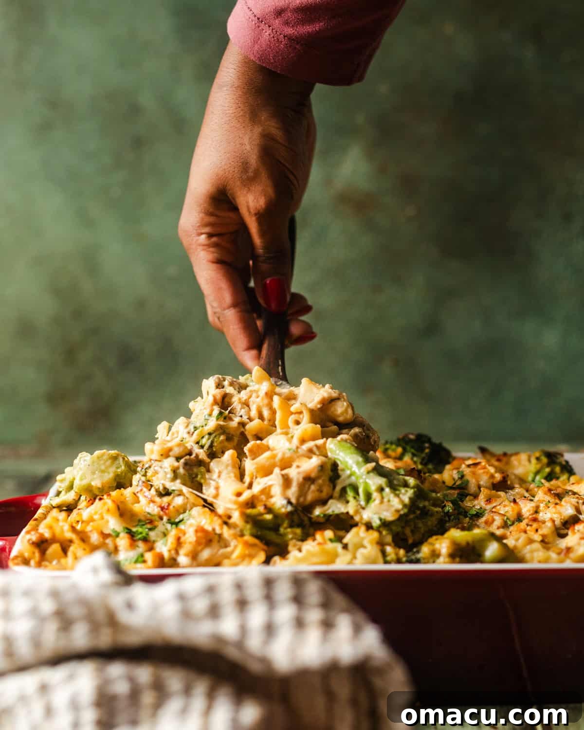 a hand scooping baked chicken and broccoli alfredo out of a baking dish. 