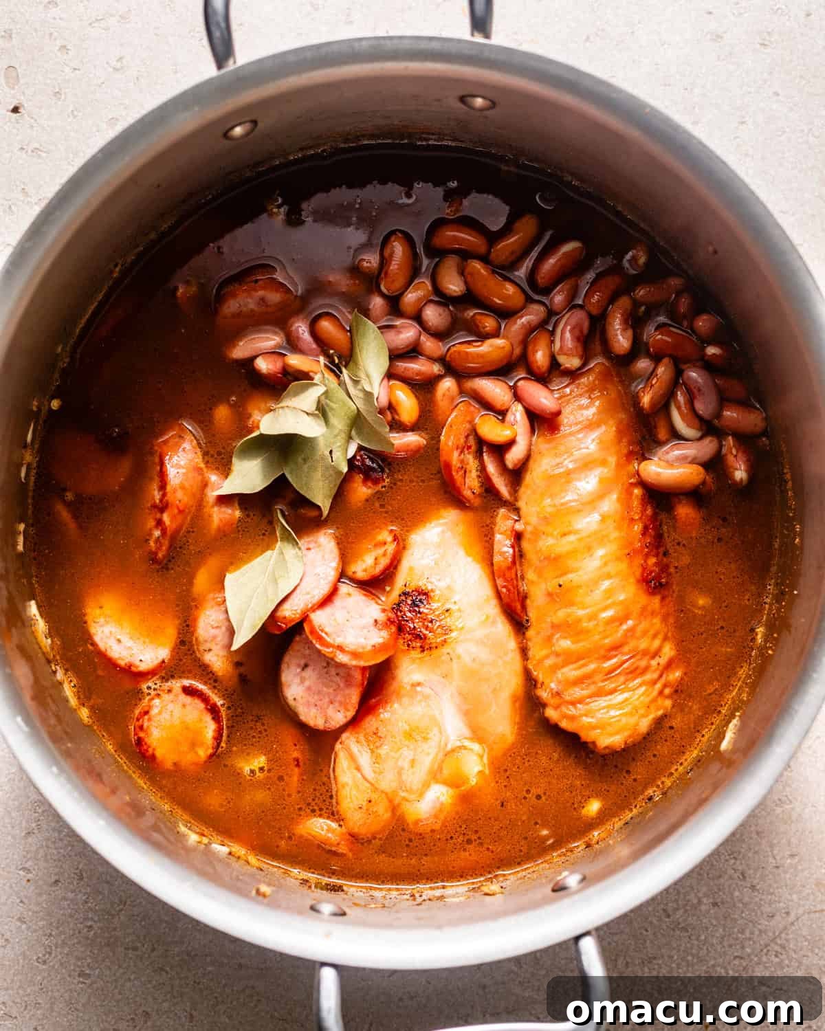 Red beans simmering in a pot with smoked turkey legs, bay leaves, fresh herbs, and browned sausage, forming a rich stew.