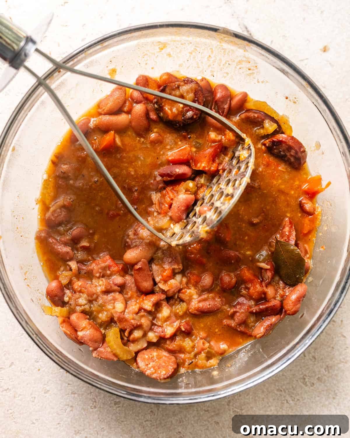 Half of the red beans being mashed in a bowl with a potato masher to create a creamier texture.