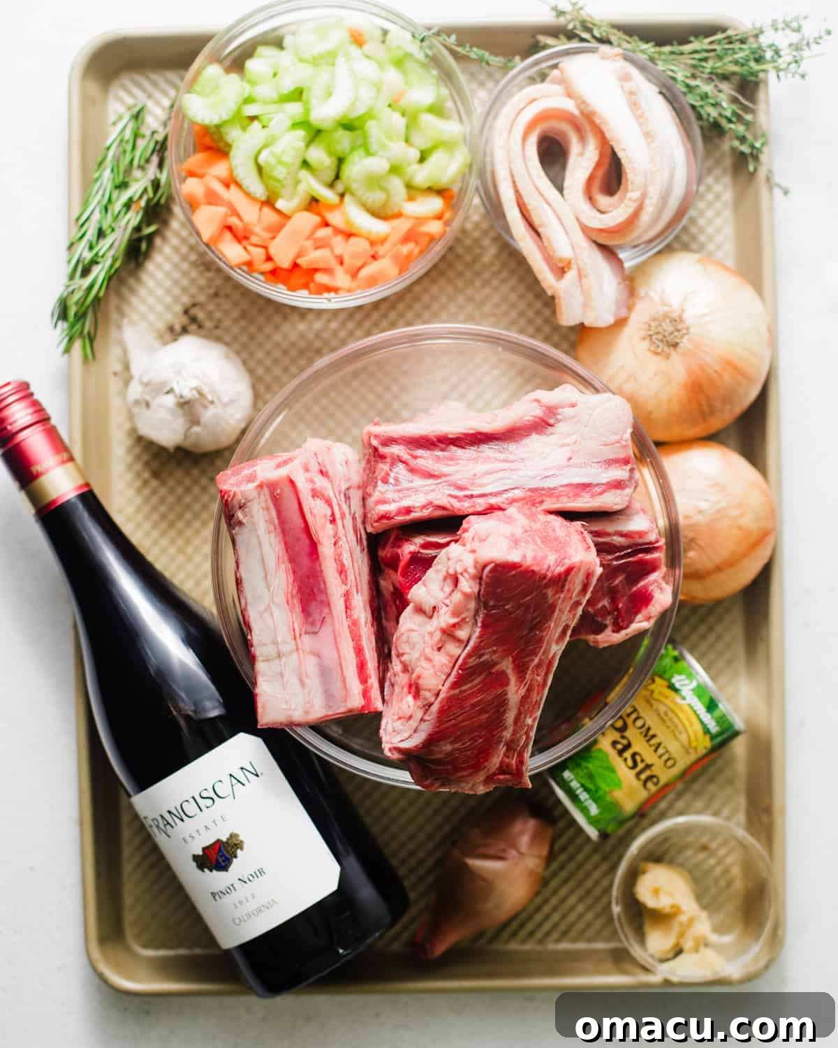 A selection of fresh ingredients laid out on a baking sheet, including beef short ribs, sliced onion, shallots, bacon strips, chopped celery, carrots, garlic cloves, tomato paste, miso paste, a bottle of red wine, heavy cream, balsamic vinegar, nutmeg, red pepper flakes, brown sugar, fresh sage, fresh thyme, fresh rosemary, bay leaves, and beef broth.
