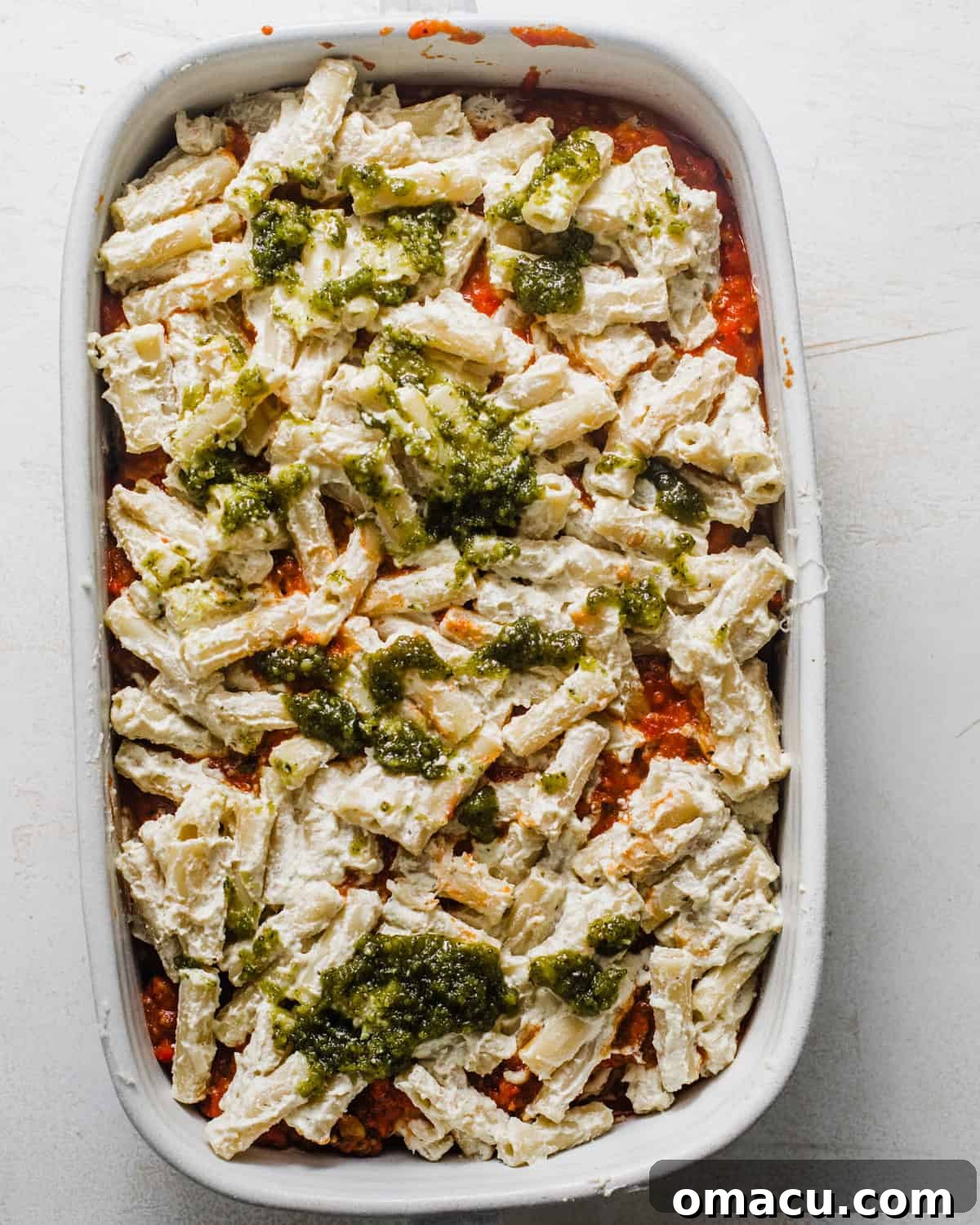 First layer of meat sauce spread at the bottom of a baking dish, topped with ziti pasta mixed with pesto and ricotta.