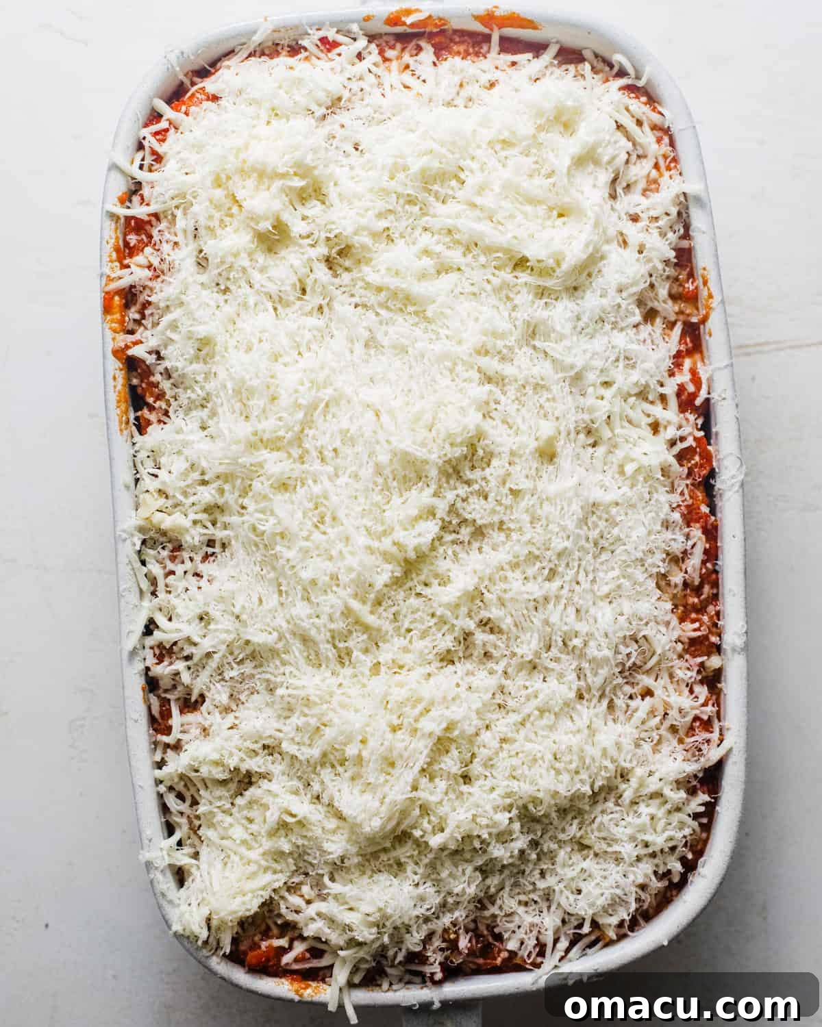 Assembled baked ziti in a baking dish, topped with cheese and dollops of pesto, ready for the oven.